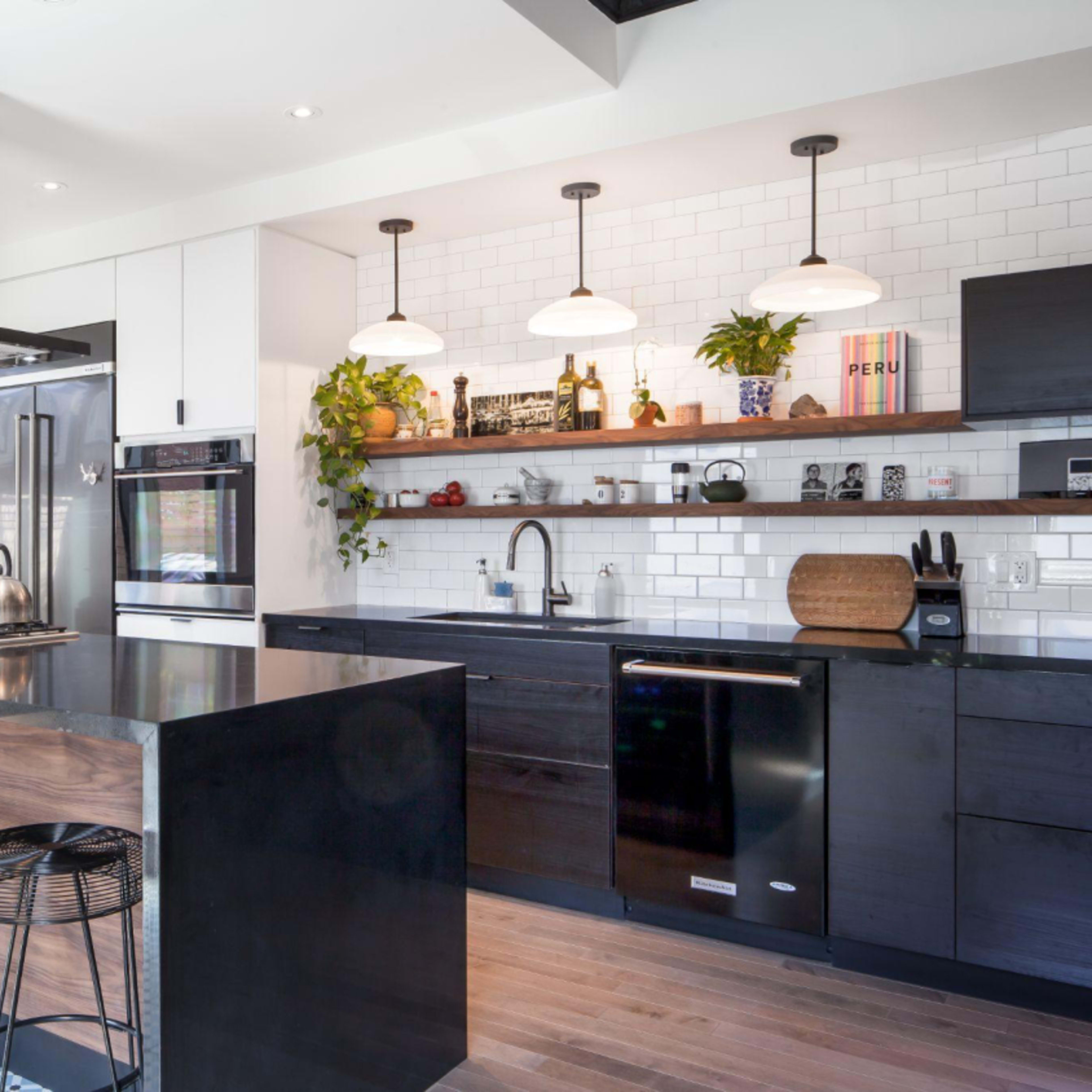 A modern kitchen featuring a large black island, stainless steel appliances, and a wall with open shelves displaying plants and decorative items.