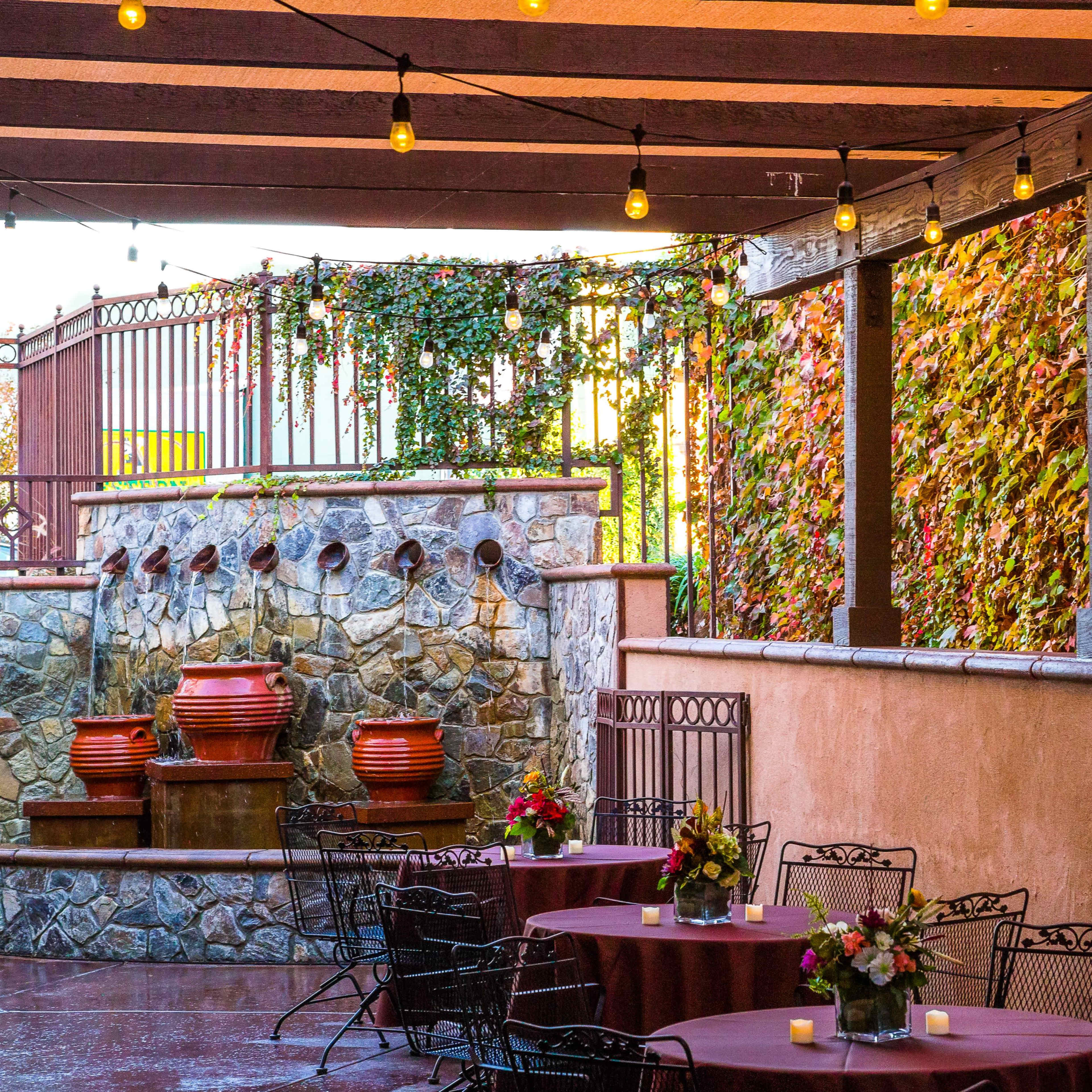 An outdoor dining area with stone walls, decorative planters, and tables set with floral centerpieces under string lights.