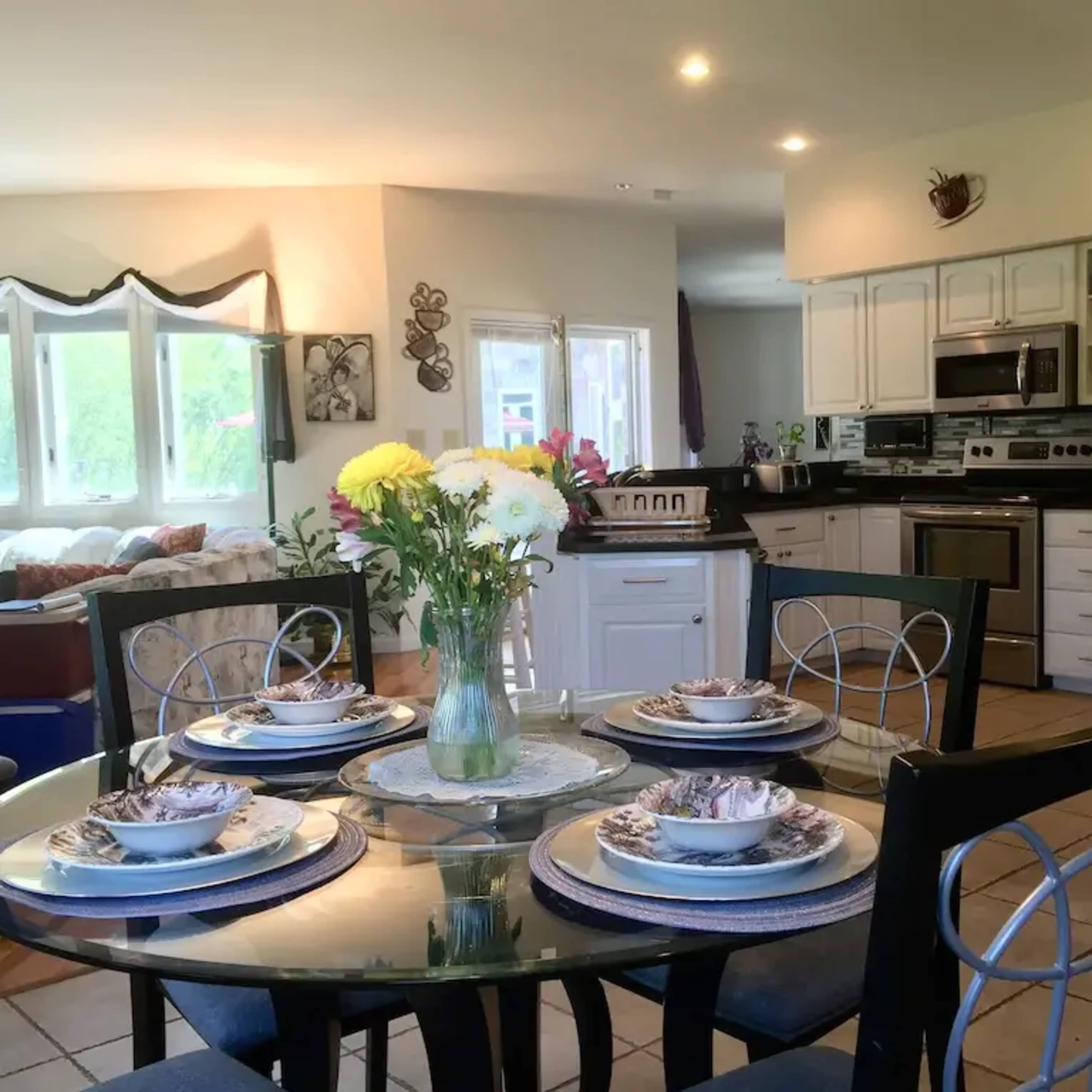A dining table with floral centerpieces and place settings is set in a modern kitchen area featuring stainless steel appliances and ample natural light.