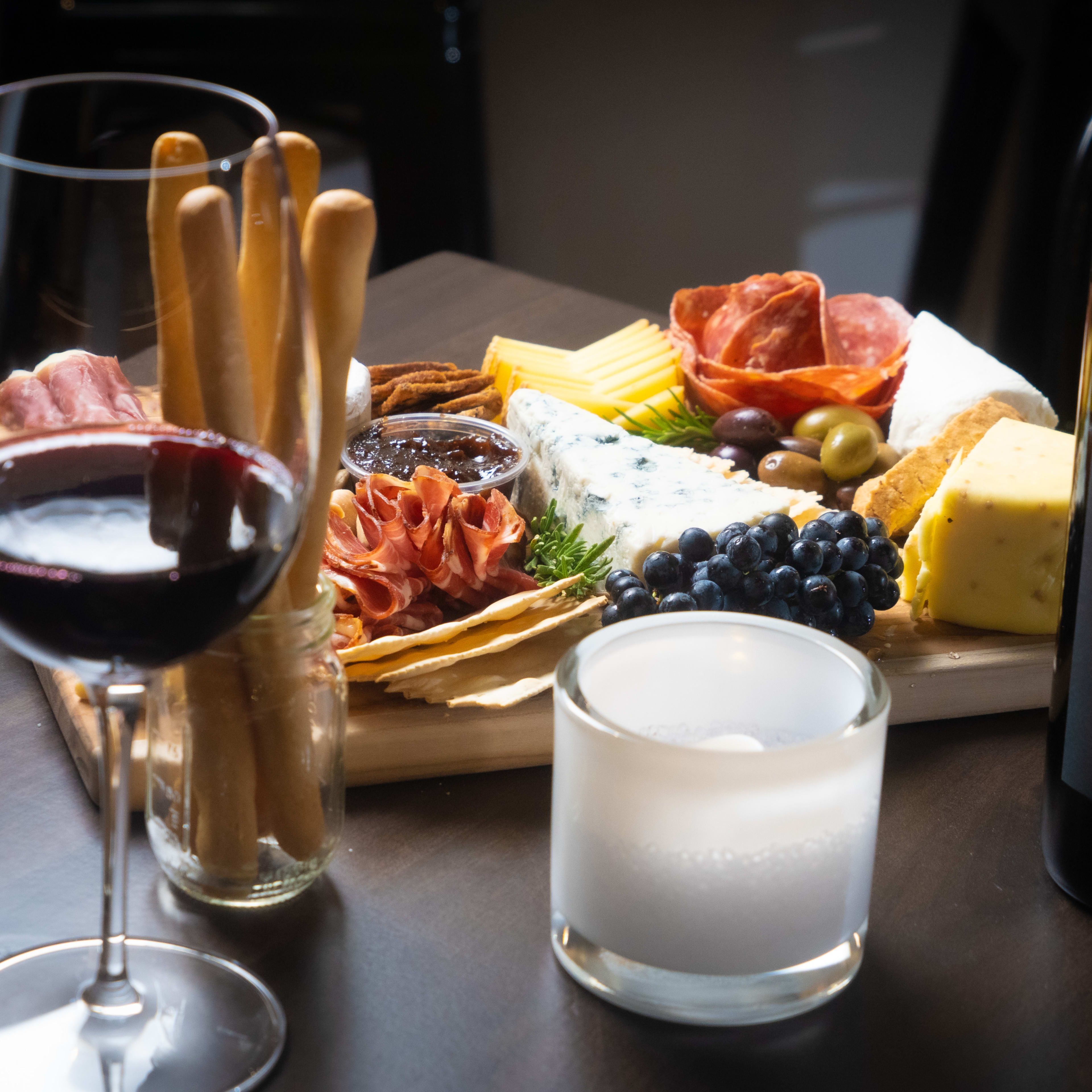 A wooden cheese board featuring various cheeses, cured meats, olives, and crackers is surrounded by two glasses of red wine and a candle.