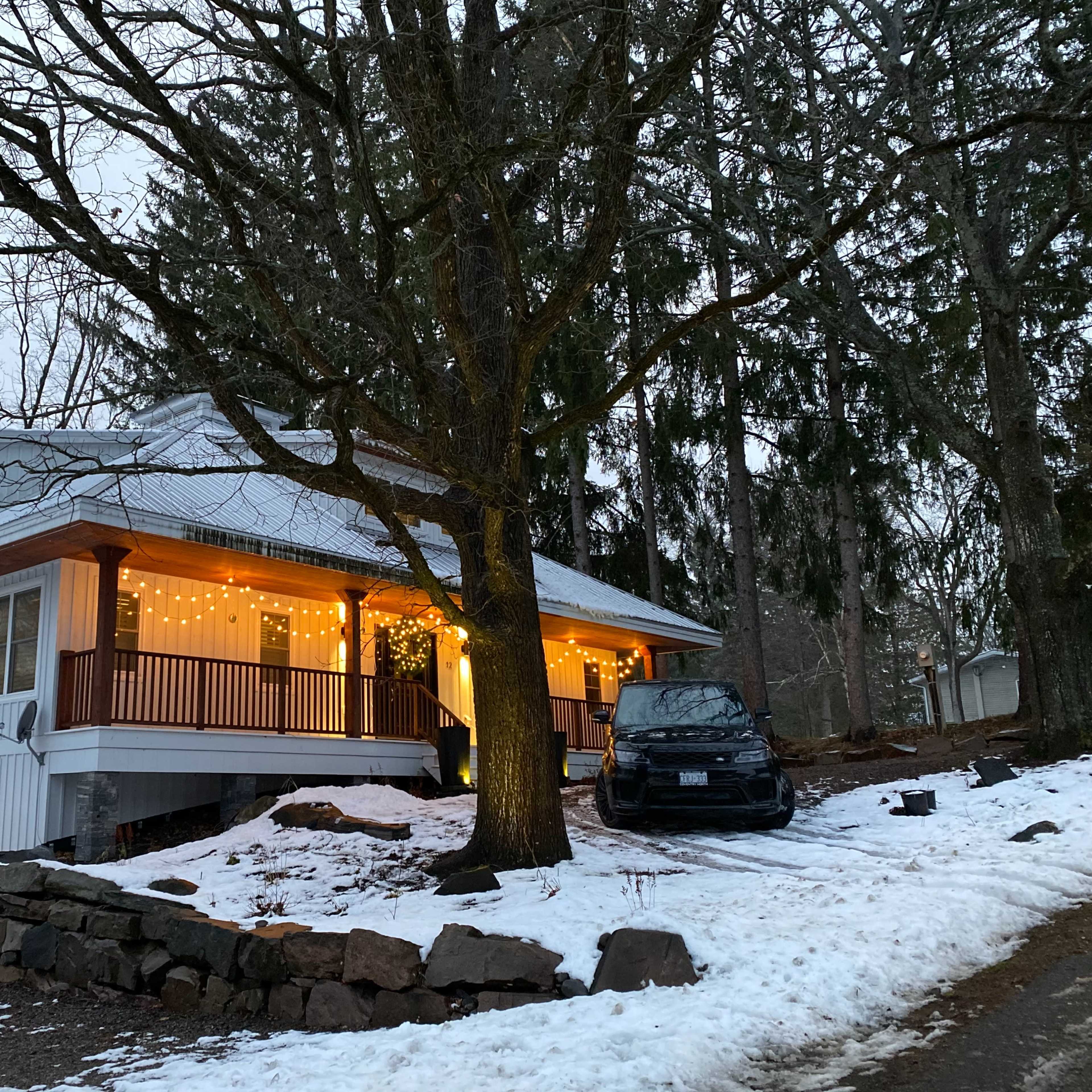 A two-story house with outdoor string lights is situated on a snowy landscape beneath leafless trees.