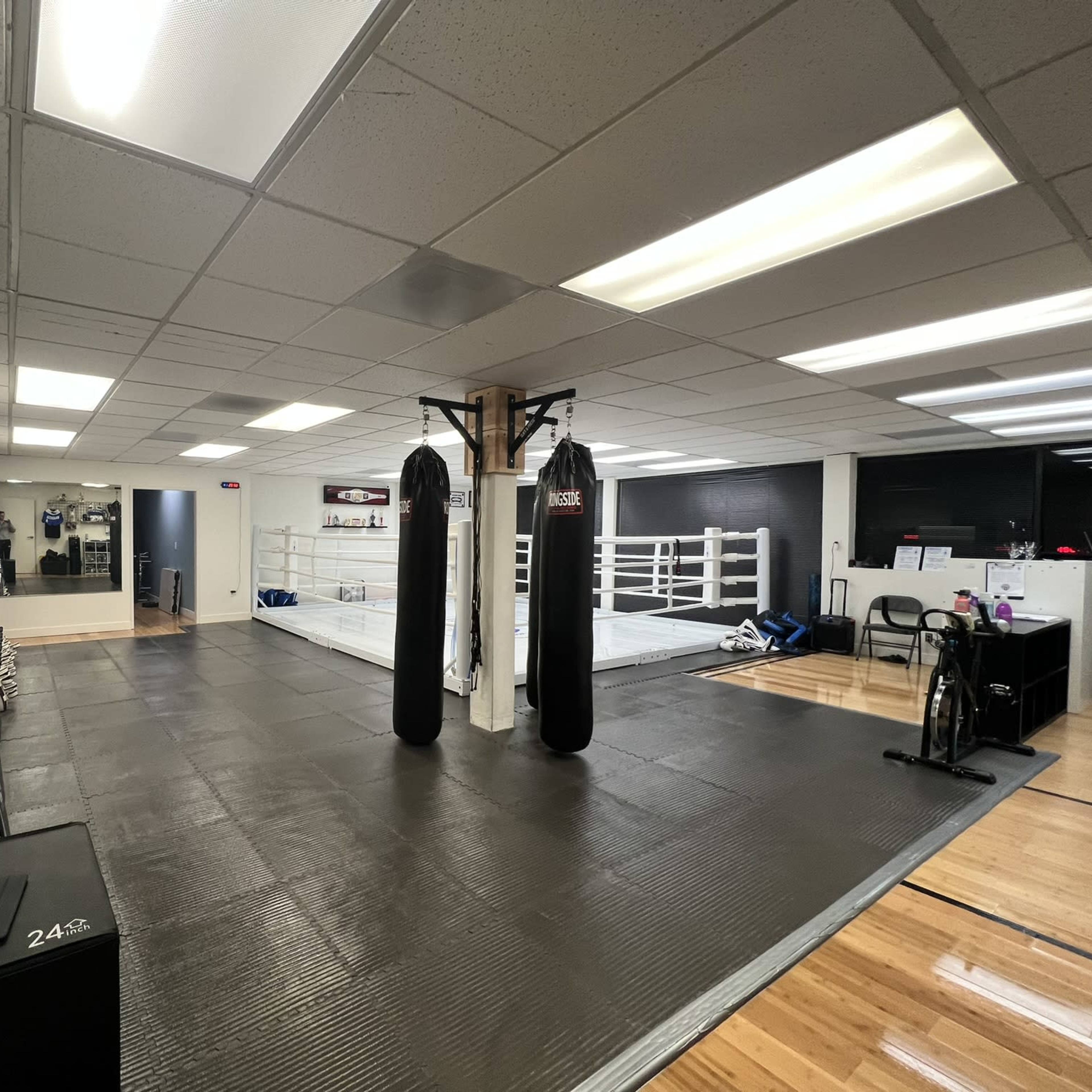 A training gym with two boxing bags, a boxing ring in the background, and several benches along the wall.