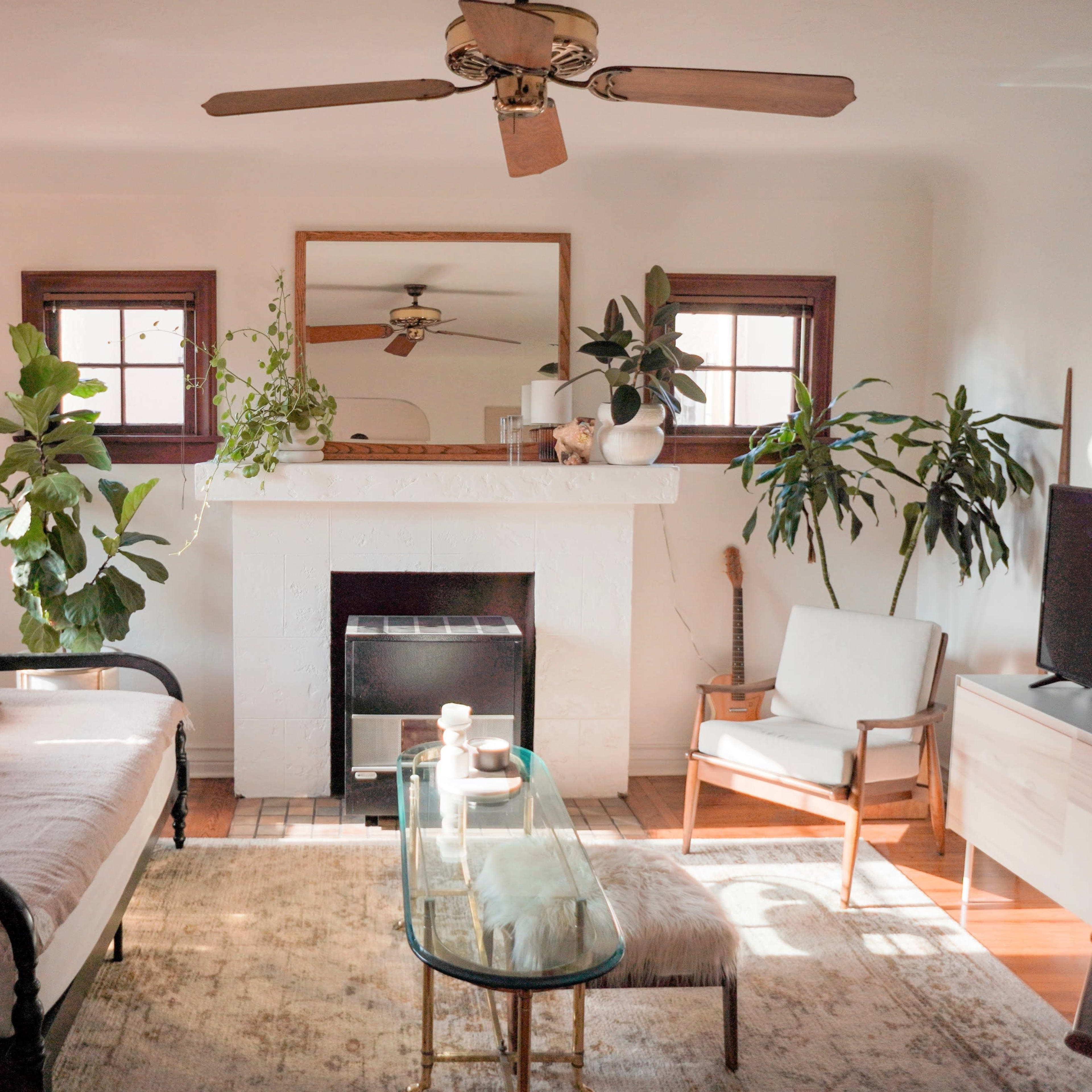 A neatly arranged living room features a sofa with decorative pillows, a glass coffee table, and plants on either side of a fireplace with a mirror above it.