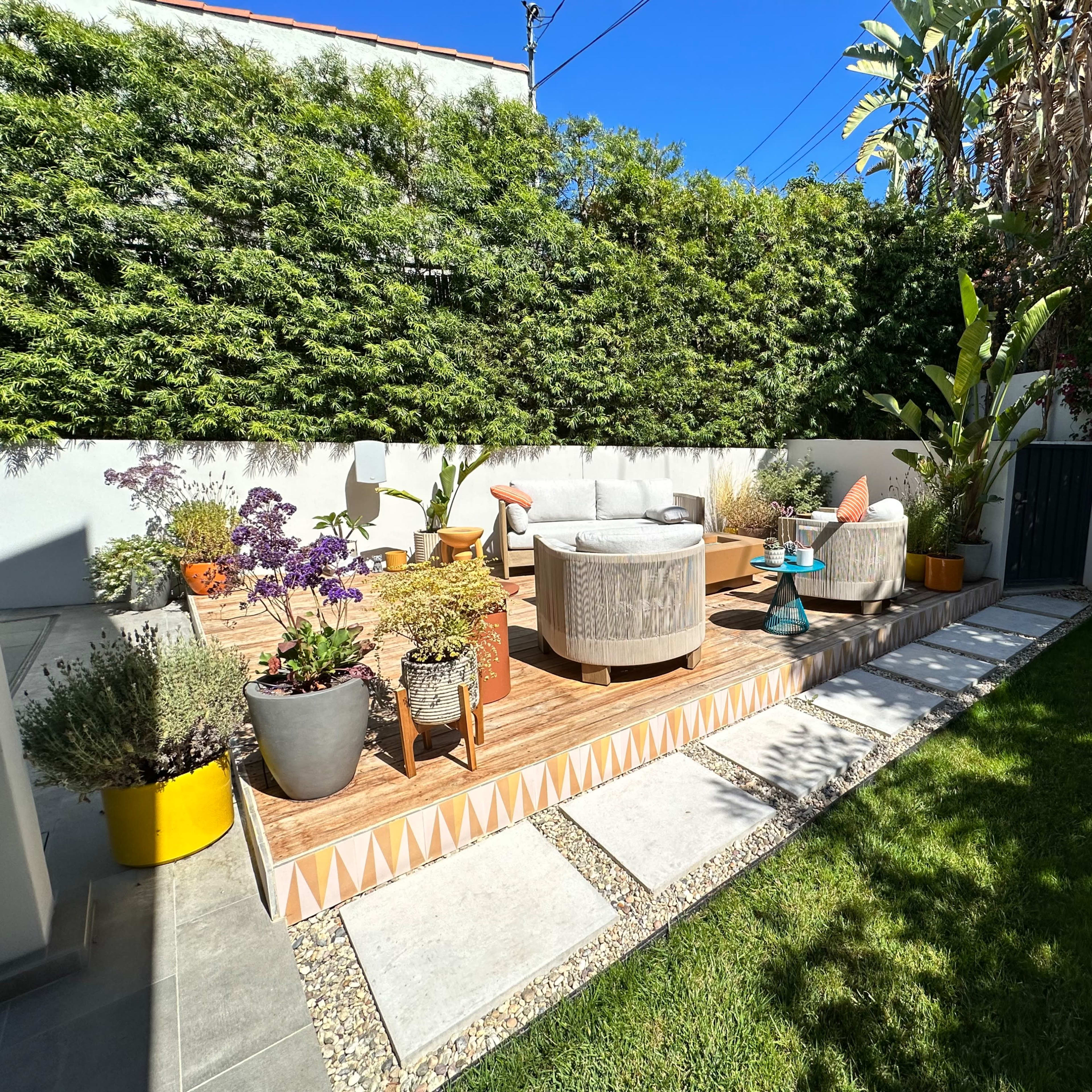 A landscaped backyard with circular seating arrangements on a wooden deck, surrounded by potted plants and a green lawn.