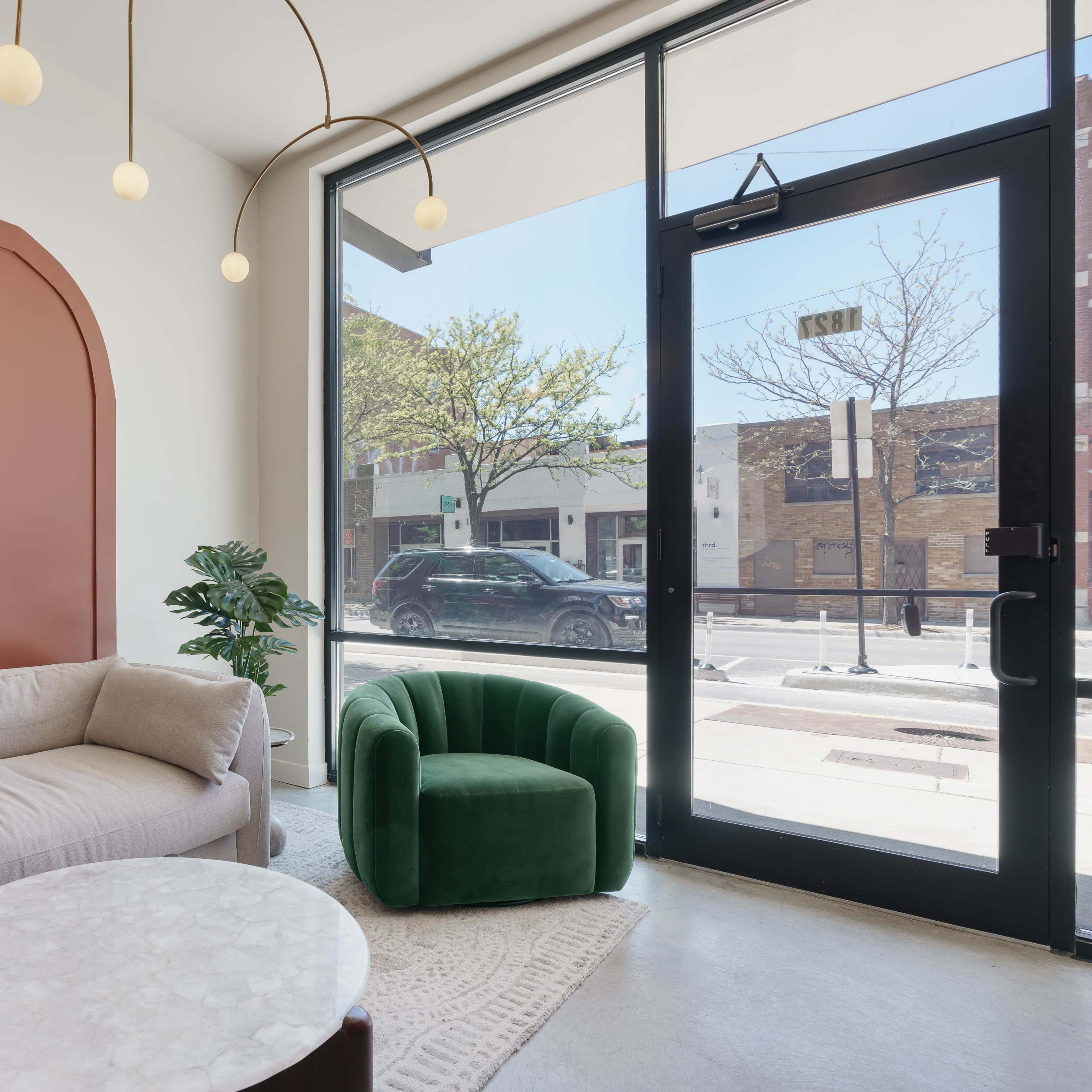 A modern interior space featuring a light-colored sofa, a green armchair, a round marble coffee table, and a large window with street views.