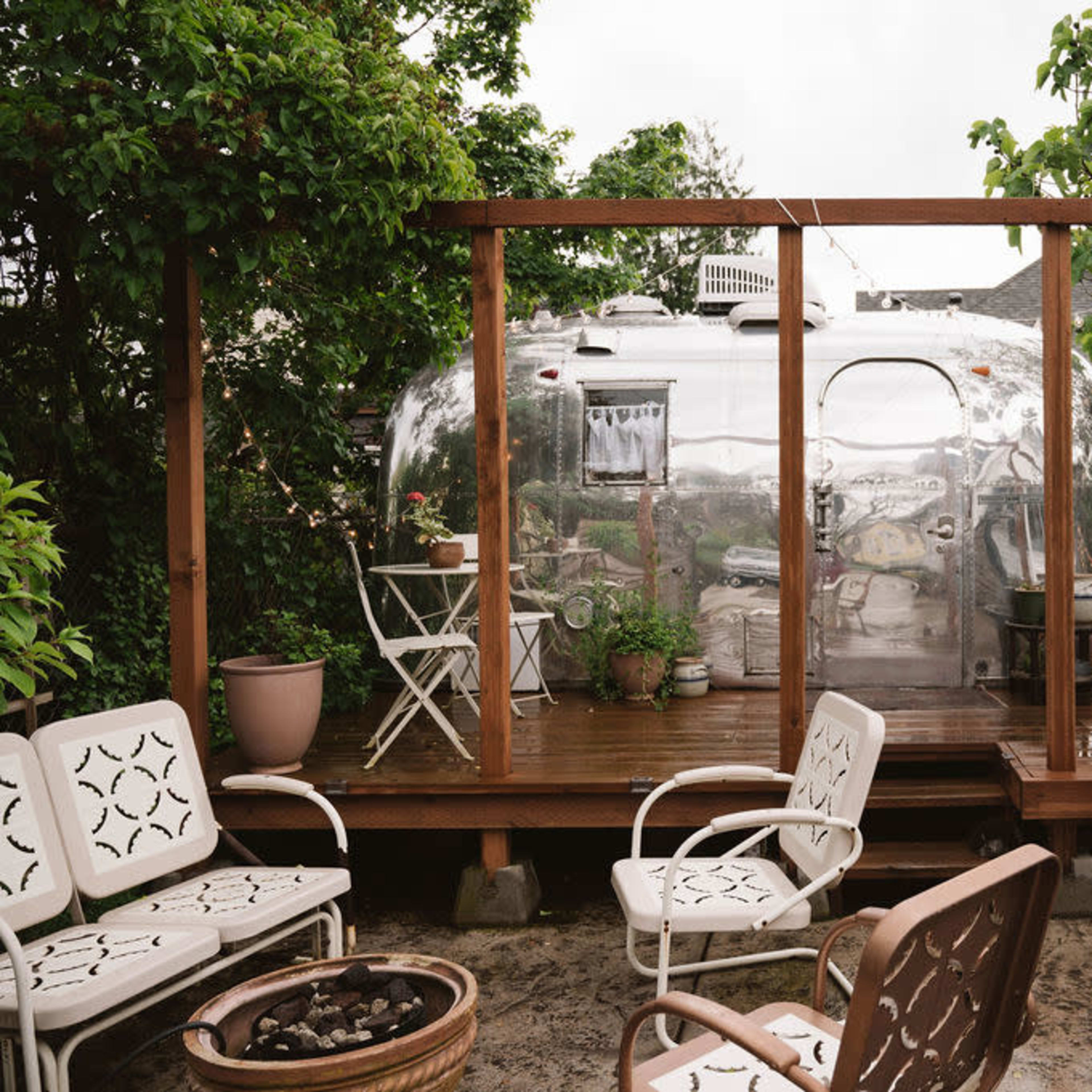 A backyard features a shiny Airstream trailer enclosed by a wooden deck surrounded by greenery and outdoor seating.