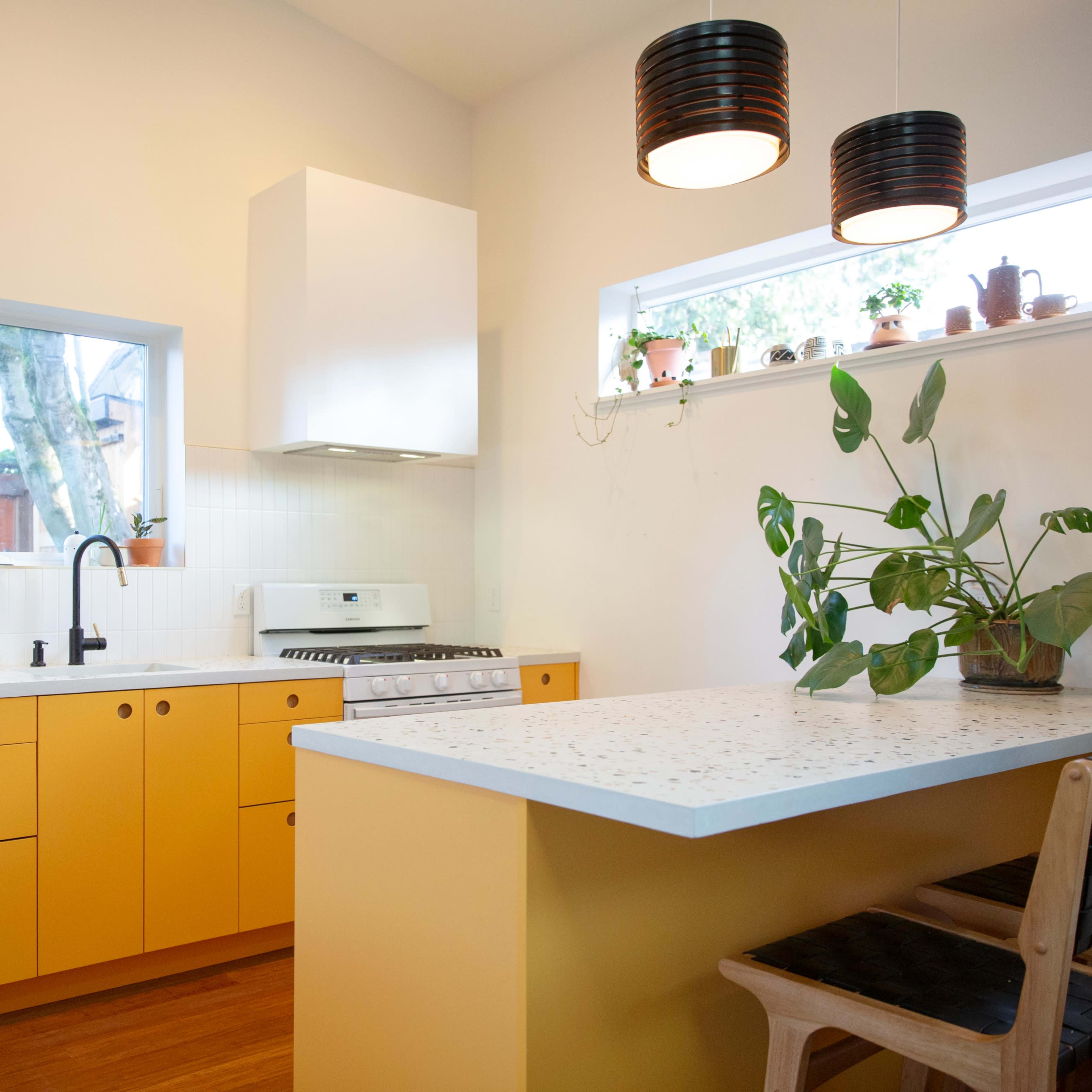 A bright kitchen features yellow cabinets, a white countertop, and a retro refrigerator, with plants decorating the windowsills and countertop.