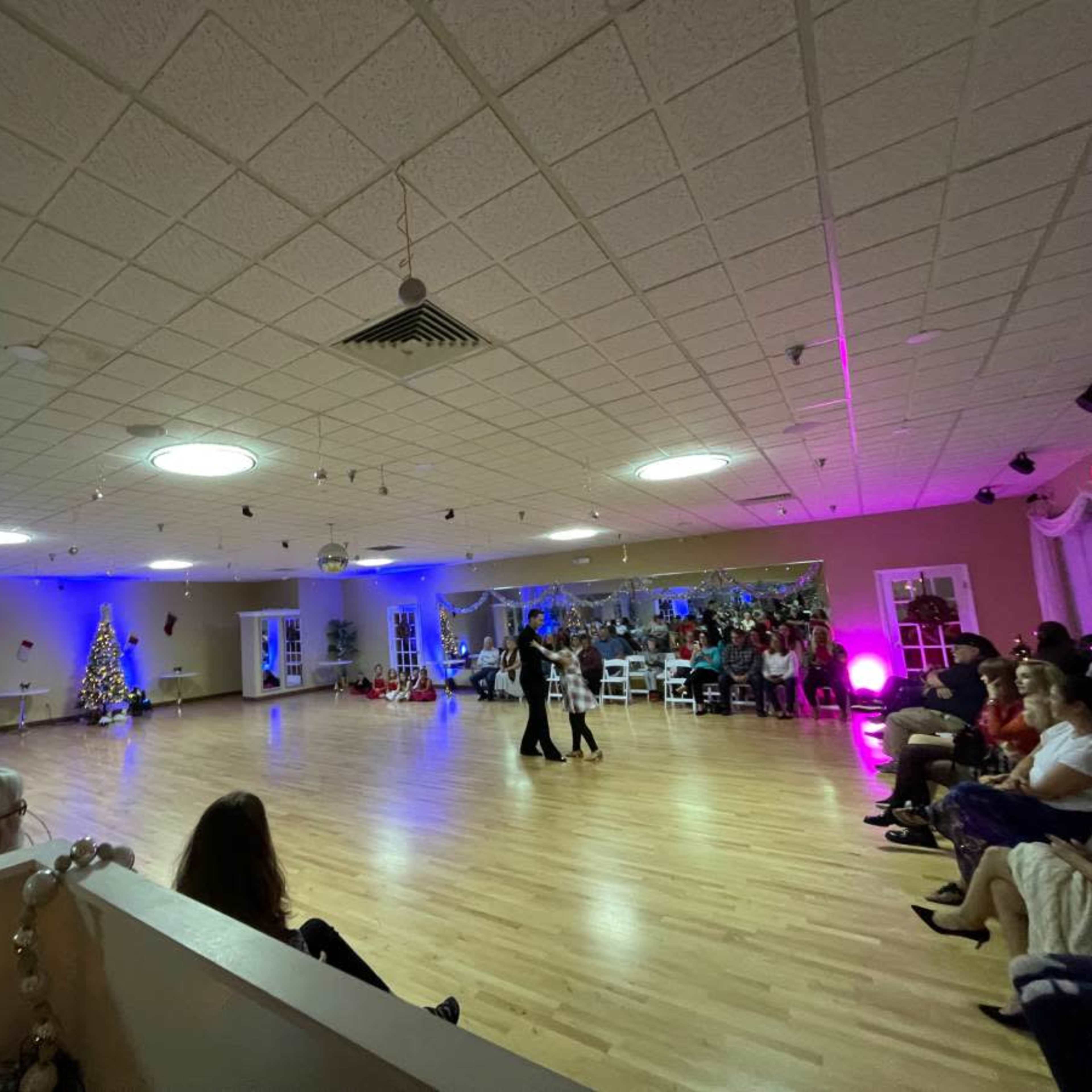 A group of people is watching a dance performance in a well-lit indoor space decorated for the holiday season.