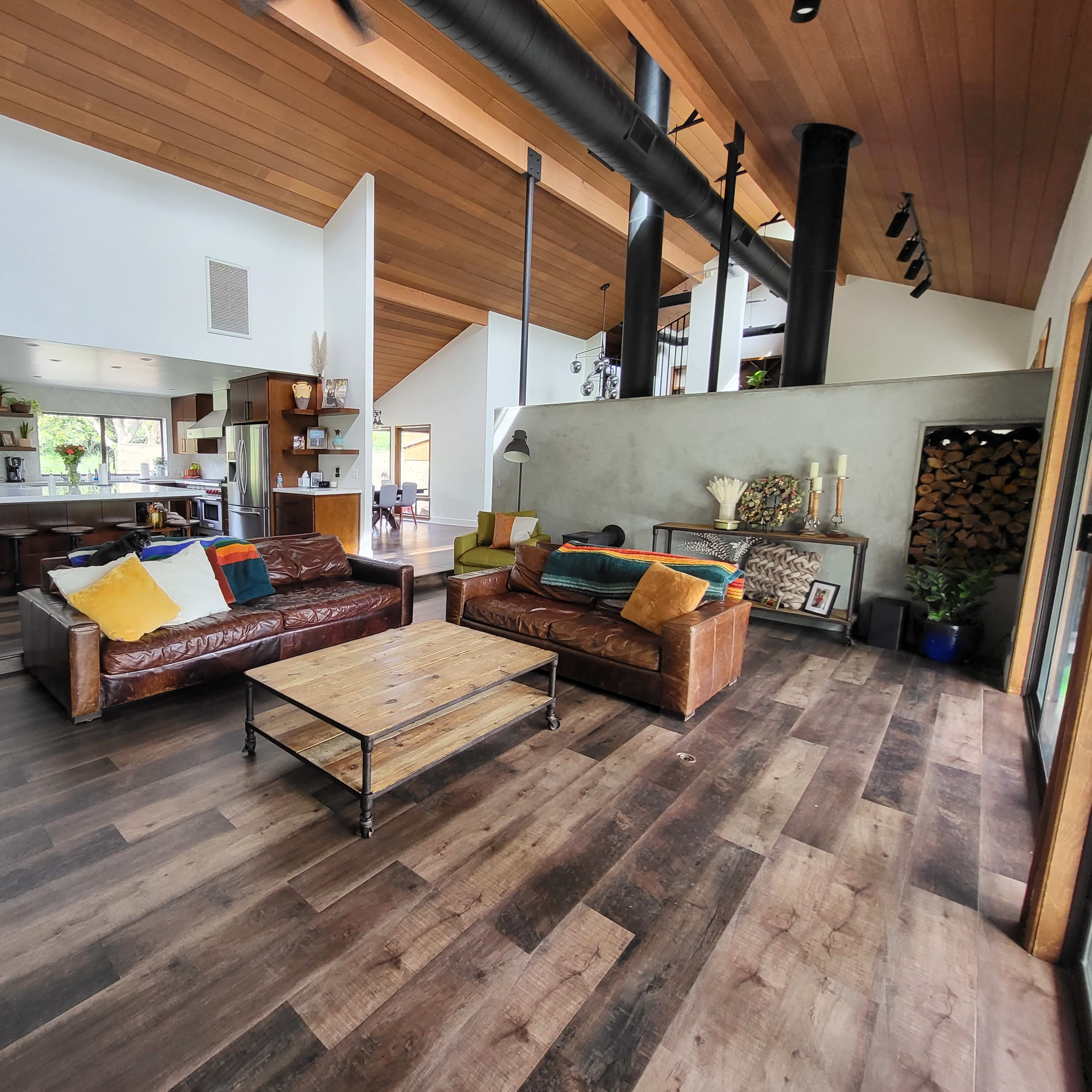 A spacious living room with wooden floors, large windows, and a modern design featuring a mix of leather and fabric sofas, a wooden coffee table, and a view of the kitchen area.