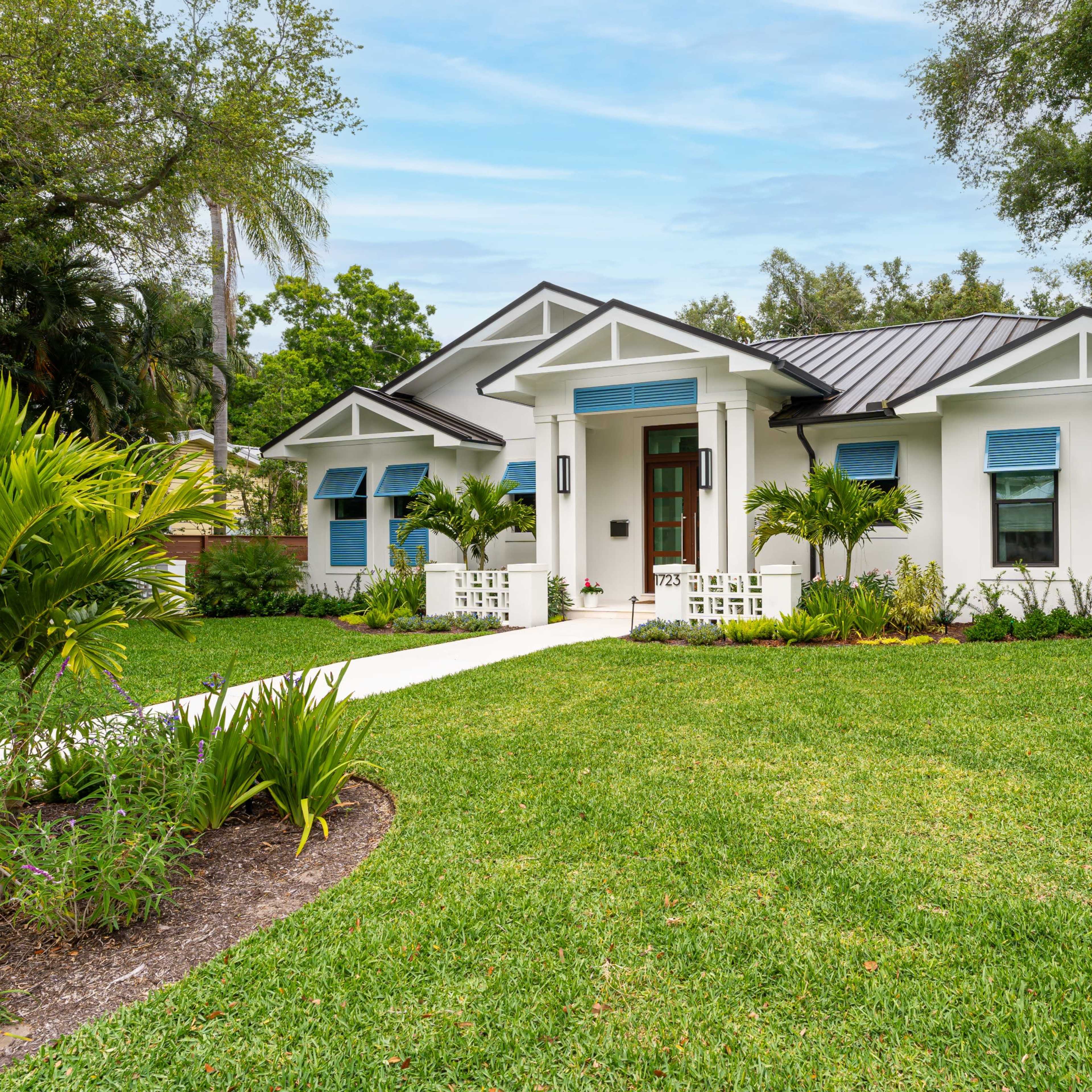 A single-story white house with a black roof, surrounded by lush greenery and well-maintained landscaping, featuring blue shutters on the windows.