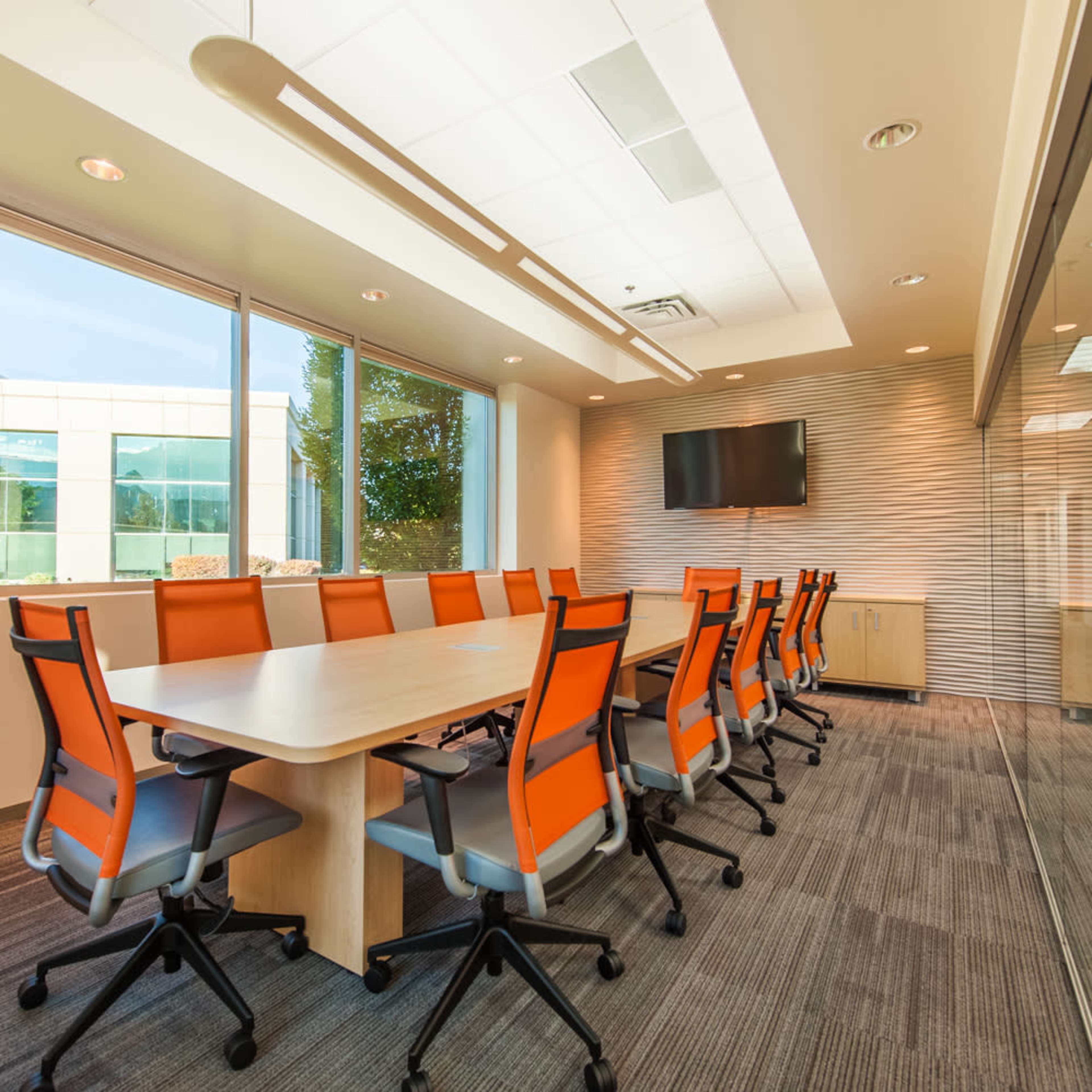 A conference room featuring a long table and orange chairs, with large windows that provide natural light and a view of the outside.