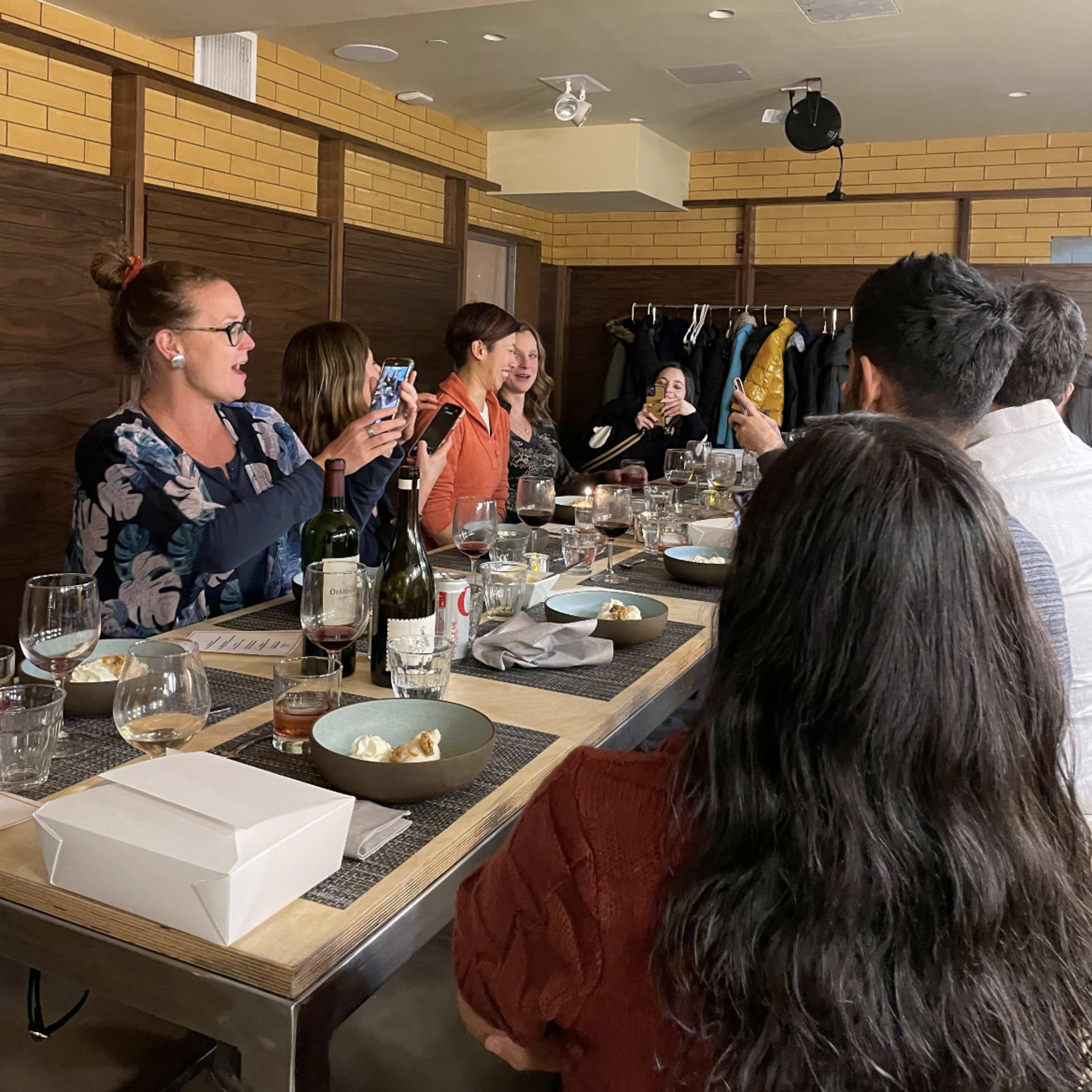 A group of people are sitting at a long table in a restaurant, engaged in conversation and enjoying food and drinks.