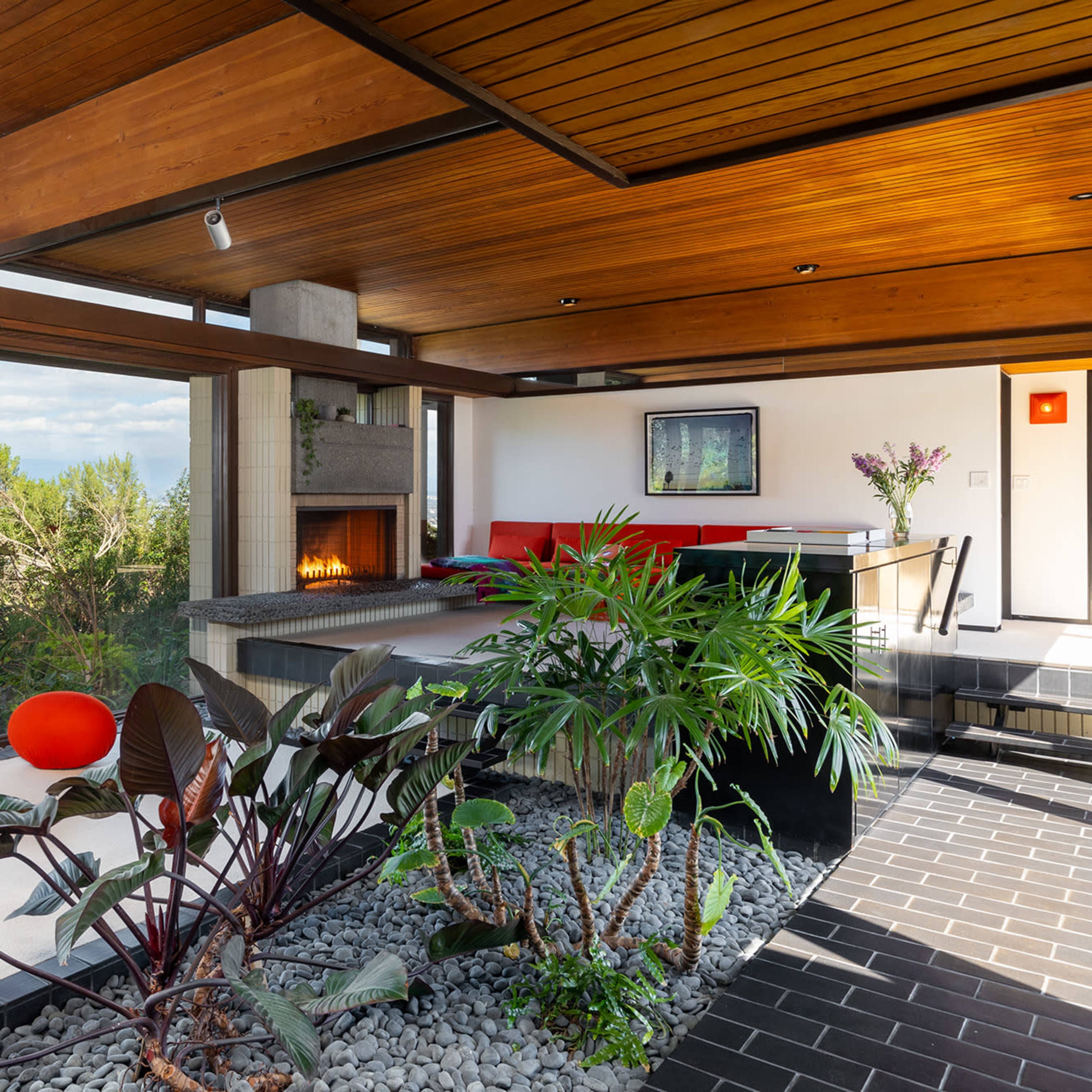 The interior of a modern home features wooden ceiling beams, a fireplace, and planters with lush greenery, alongside contemporary seating areas.