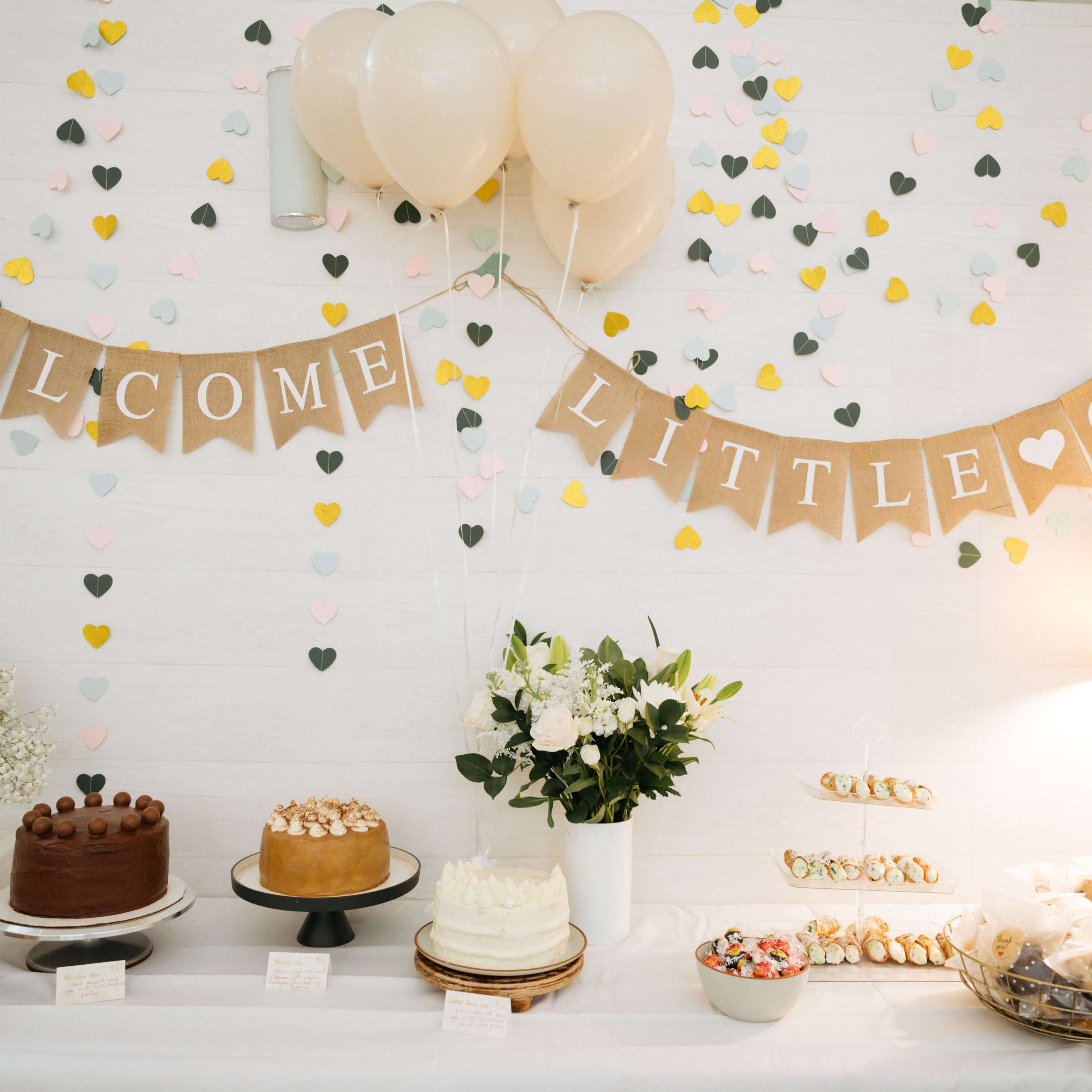 A dessert table is decorated with a "Welcome Little One" banner, balloons, and an assortment of cakes, cookies, and flowers.