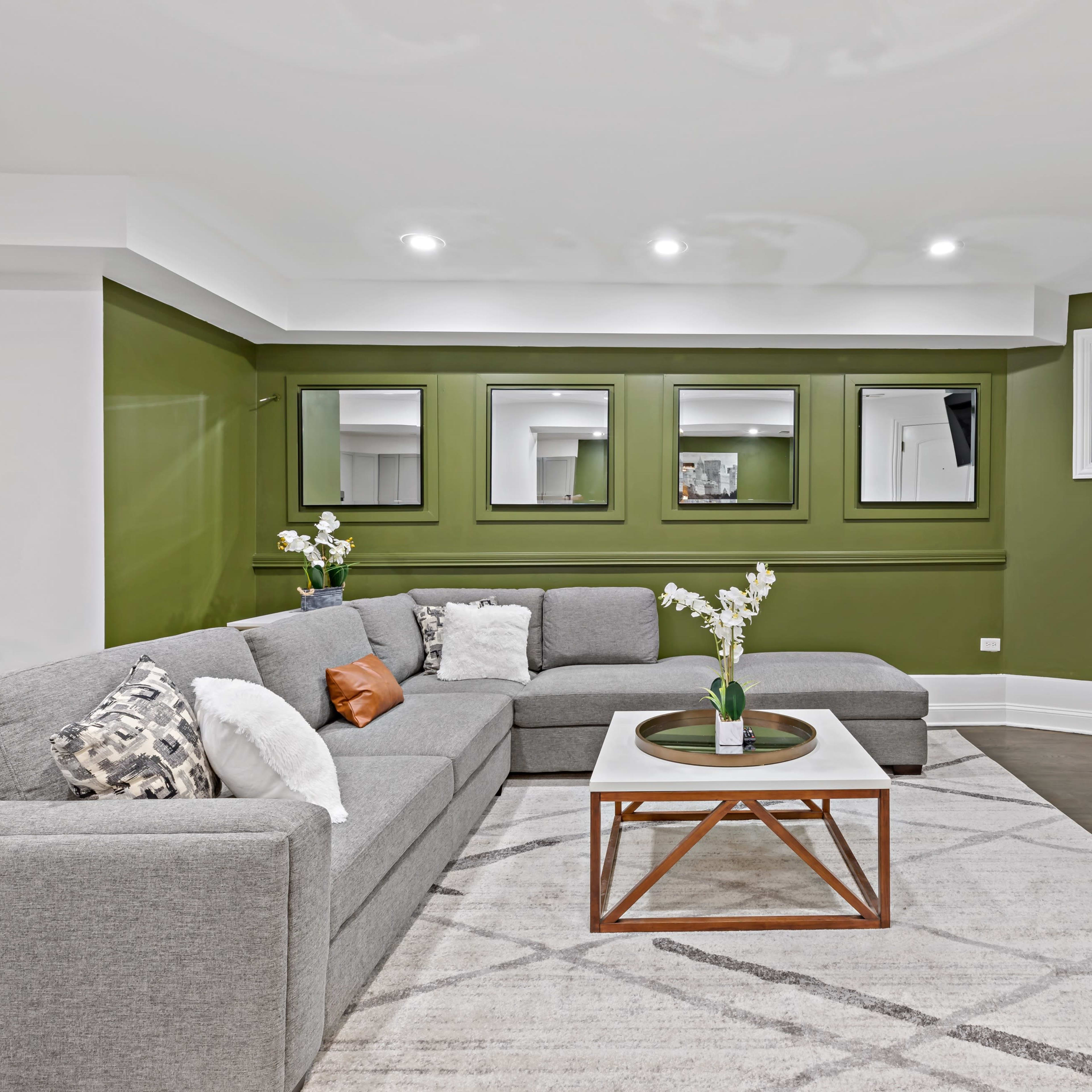 The living room features a sectional gray sofa, a white coffee table with a wooden base, a fireplace, and green walls with mirrors and plants.