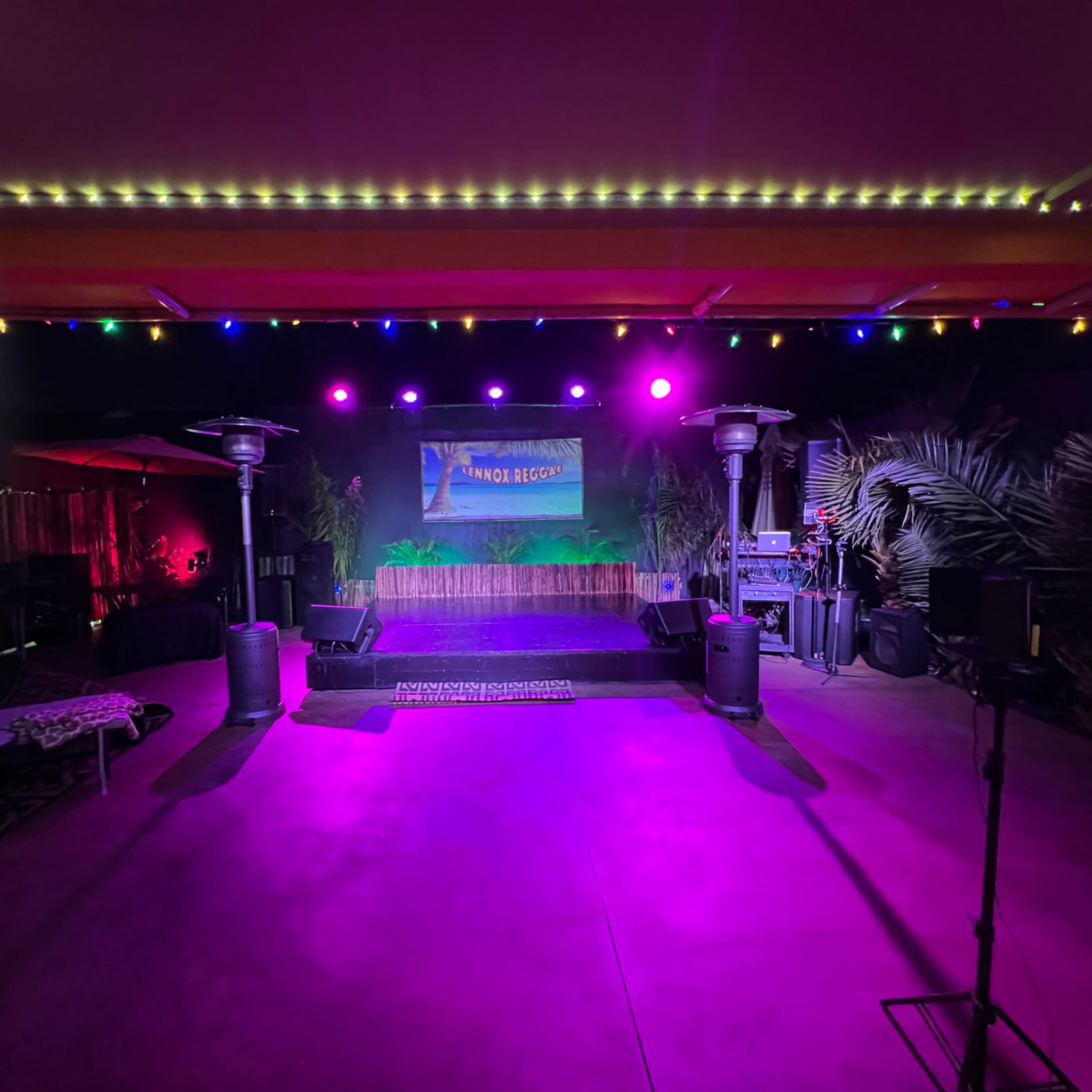 A stage set up outdoors with colorful lights, surrounded by decorative plants and equipment for a performance or event.