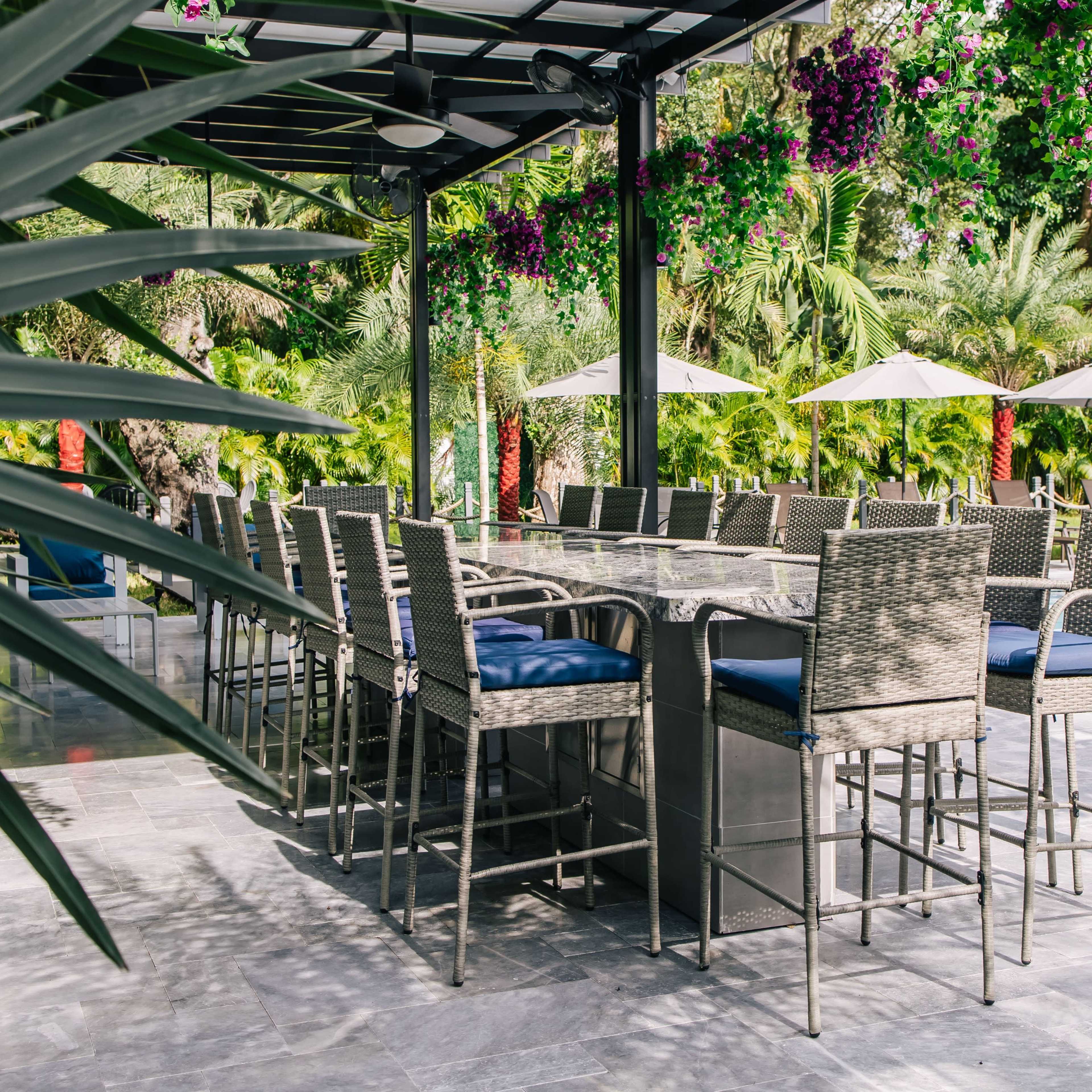 A poolside outdoor seating area features high chairs and a large table under a pergola adorned with hanging flowers.