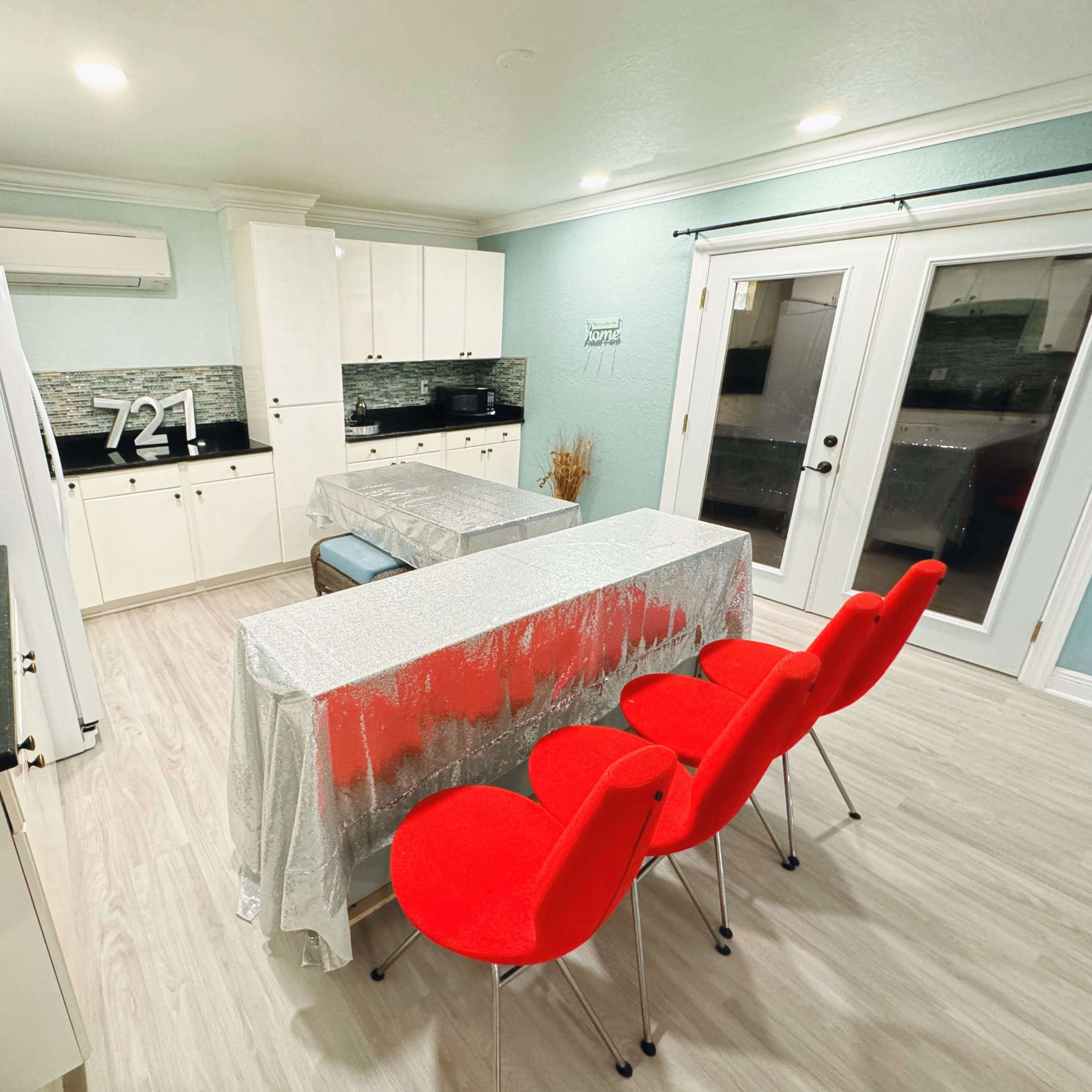 A modern kitchen features a black countertop, white cabinets, and a dining table covered with a silver tablecloth, accompanied by red chairs.
