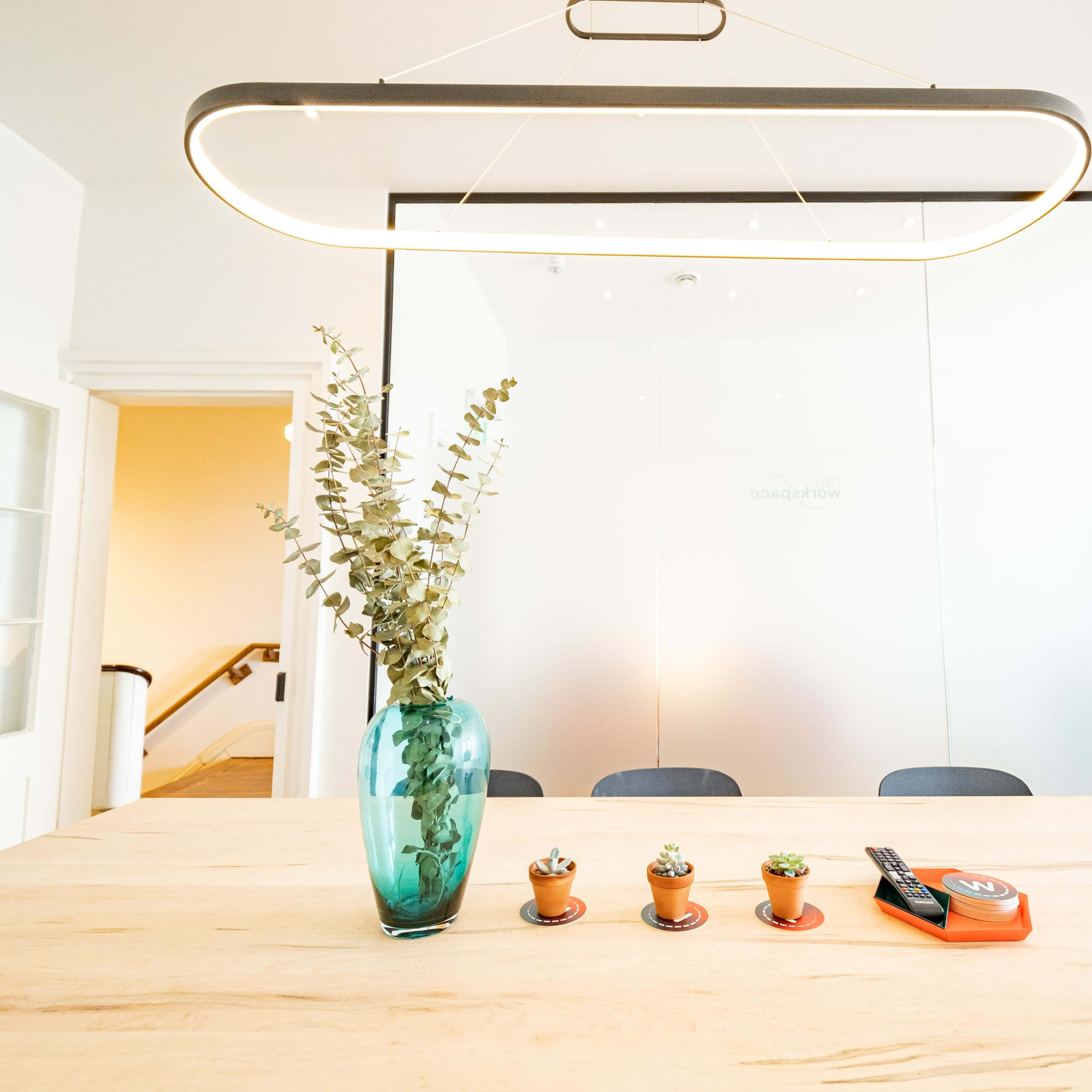 A modern conference room features a wooden table with a turquoise vase, small potted plants, and a measuring tape under a hanging light fixture.