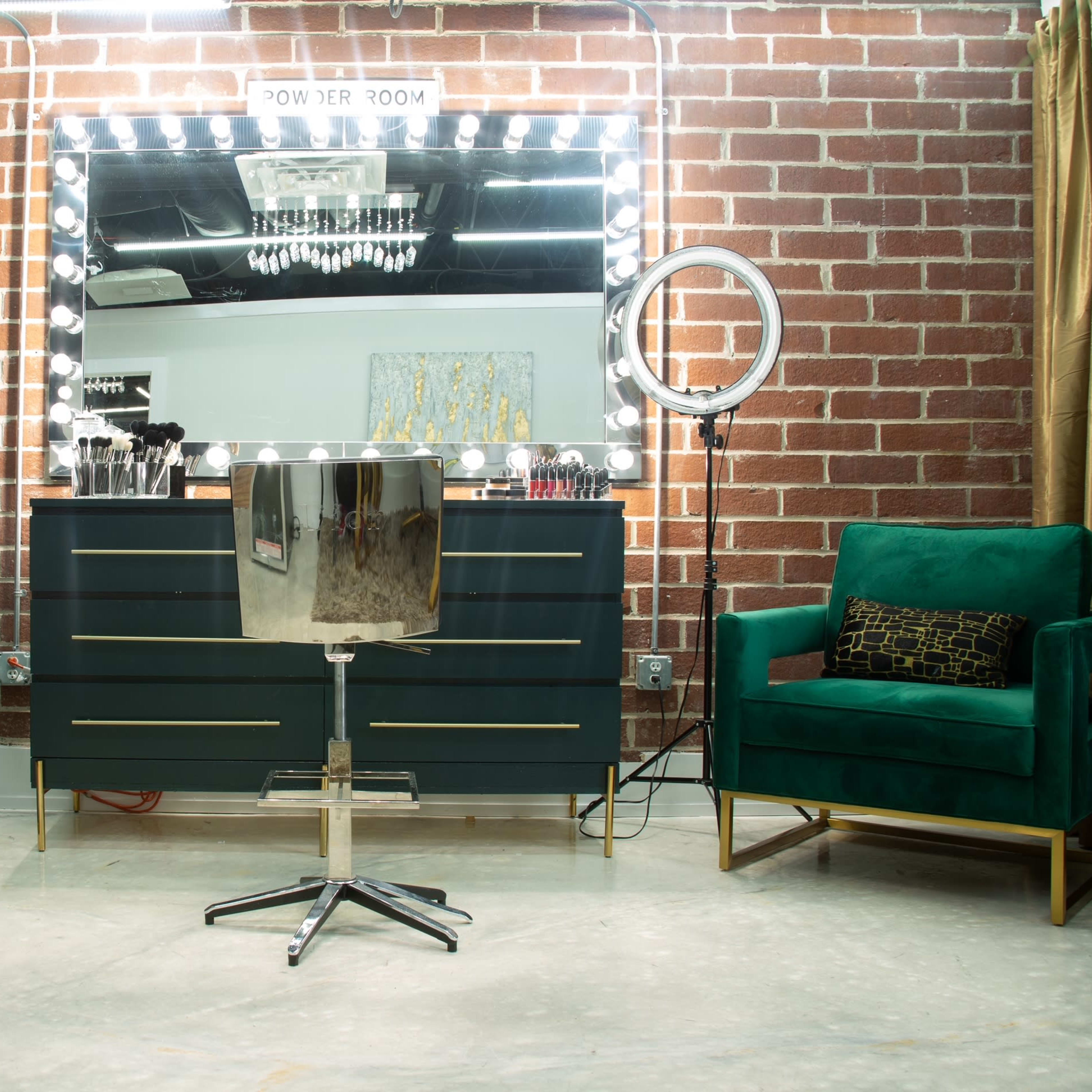 A stylish beauty setup featuring a large mirror with lights above it, a metallic salon chair in front, and two green velvet armchairs on either side, all set against a brick wall.