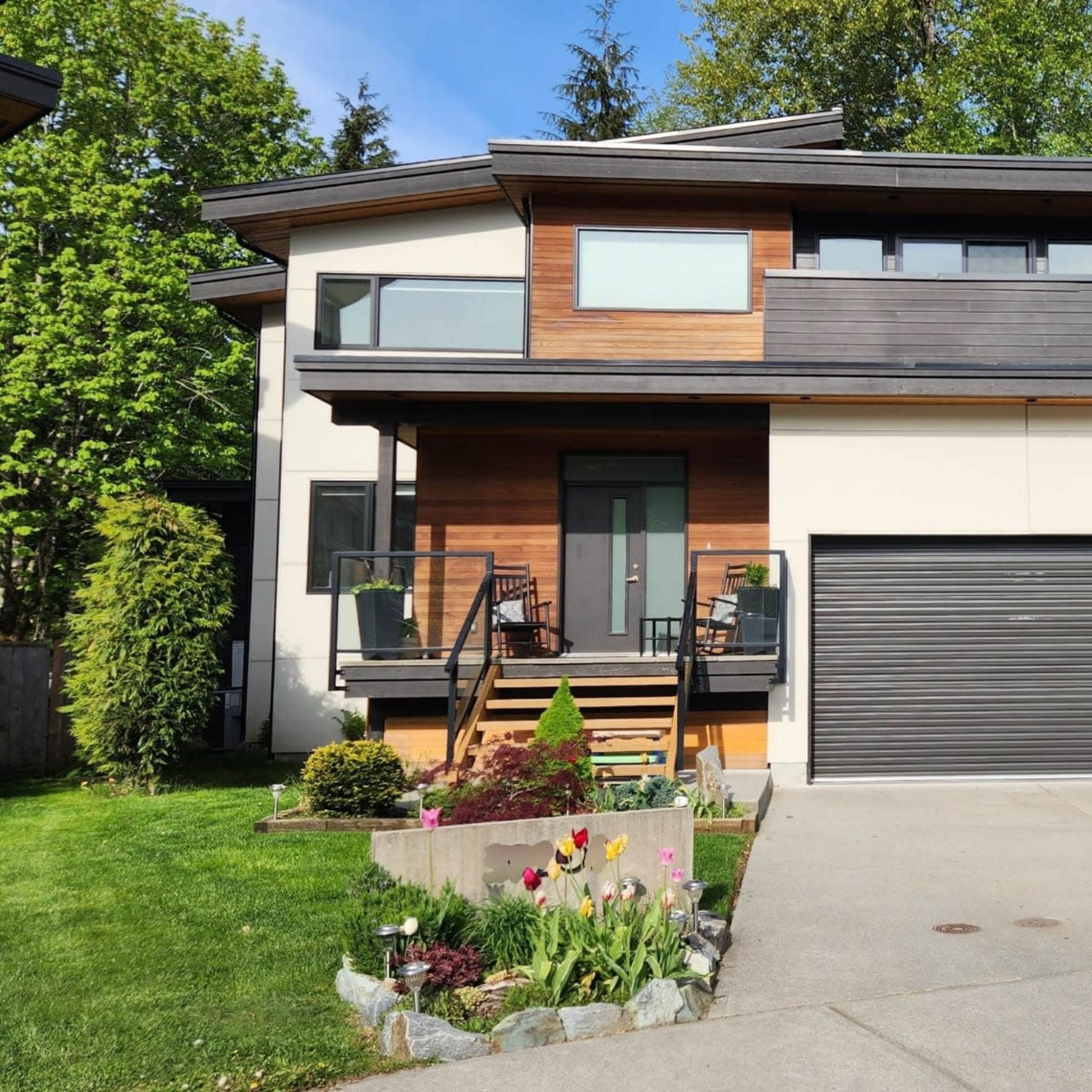 A modern two-story house with a wooden facade and a landscaped front yard featuring flowers and a paved driveway.