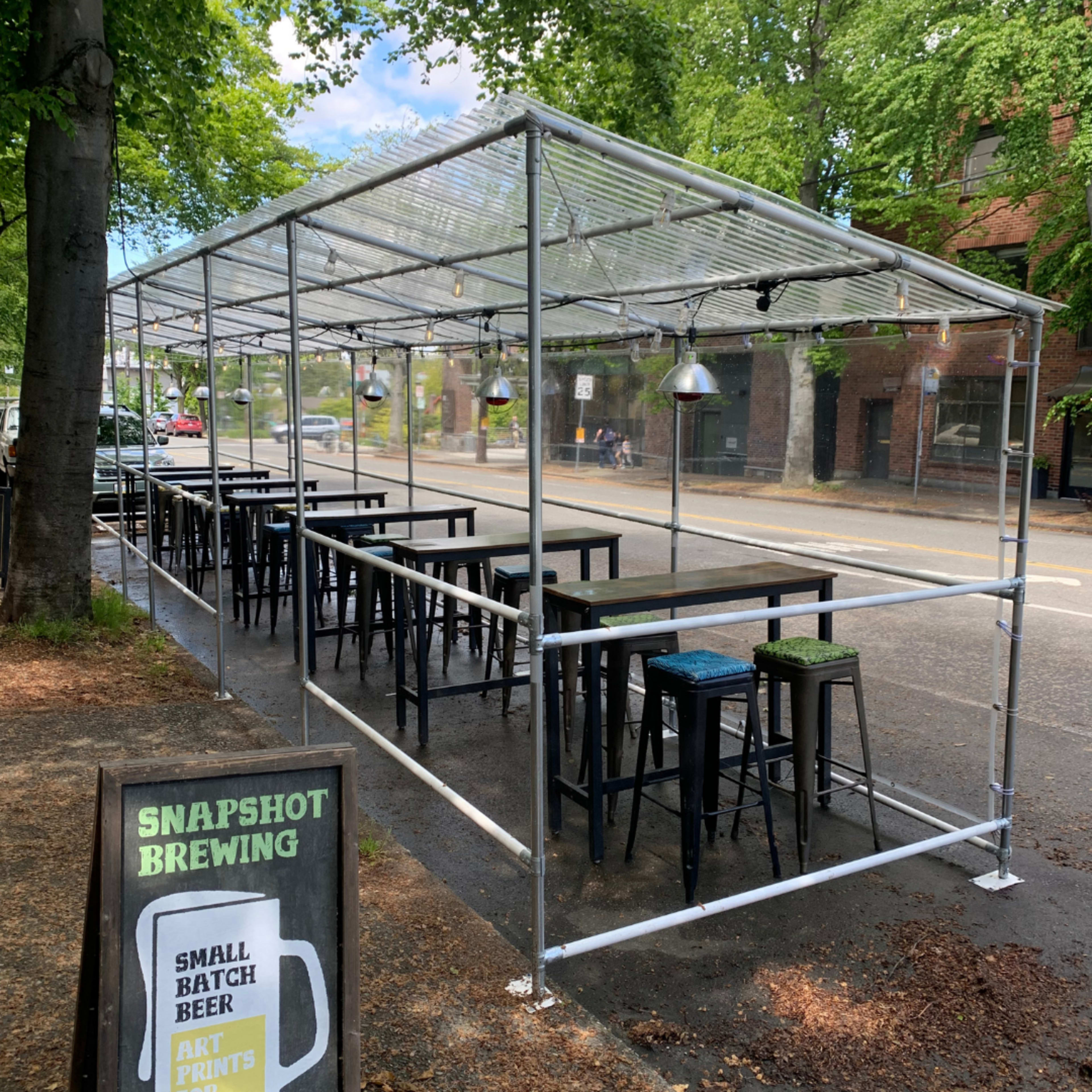 A clear, tent-like structure with black tables and stools is set up on a street outside Snapshot Brewing, surrounded by trees and a sign advertising small batch beer.