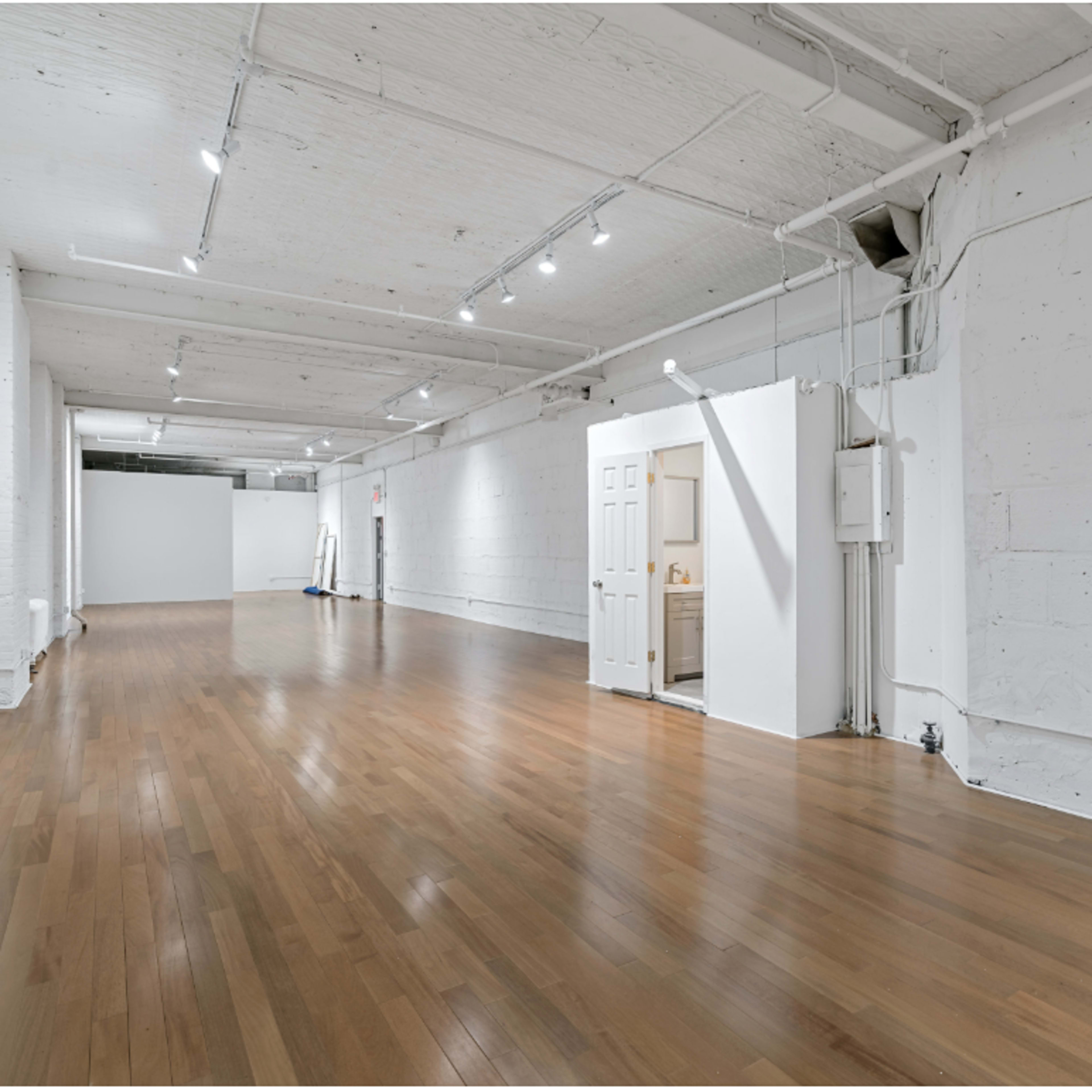 A spacious, empty interior room with wooden floors, white walls, and several overhead lights.