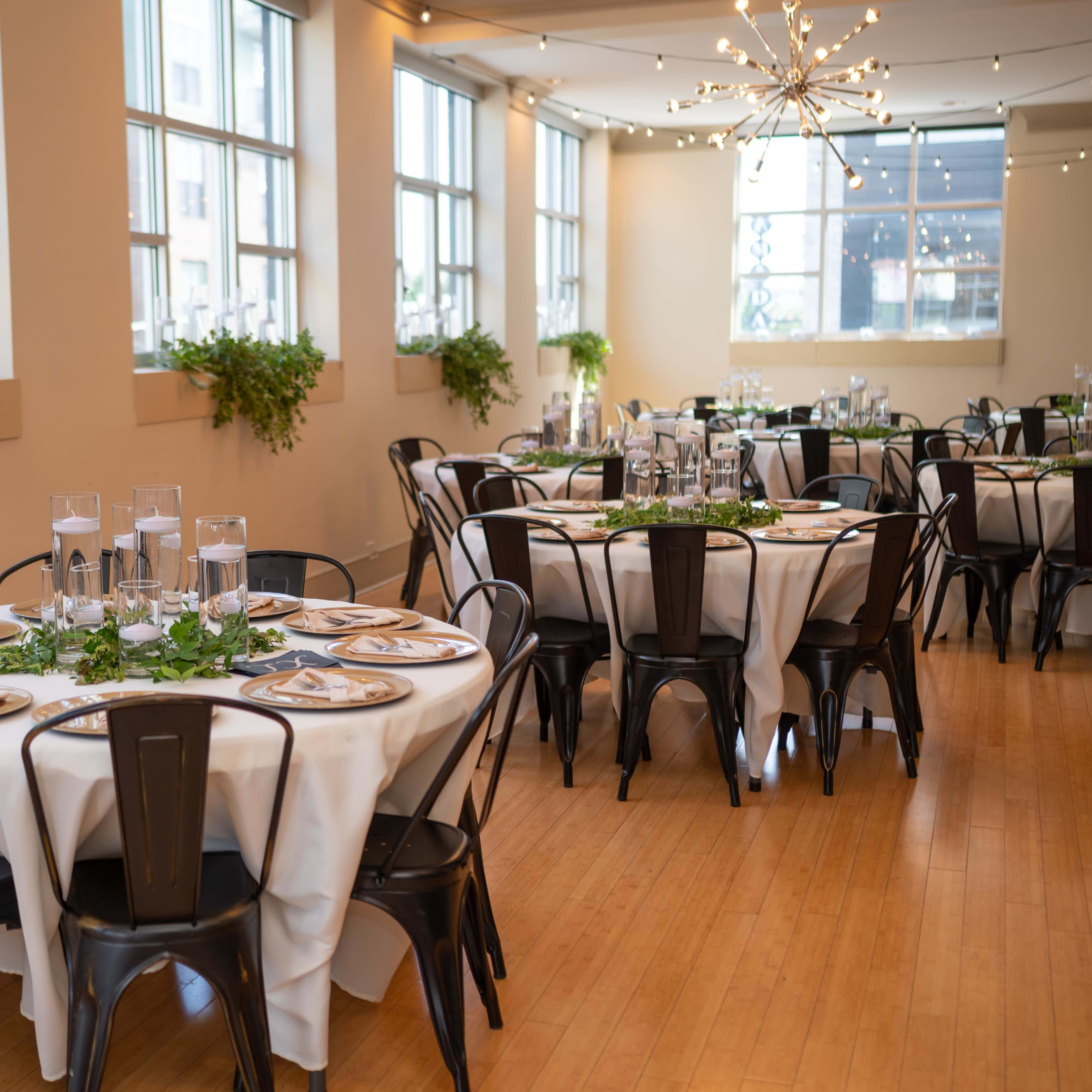 A banquet hall features round tables set with white tablecloths and black chairs, decorated with green foliage and glass centerpieces, illuminated by natural light through large windows.