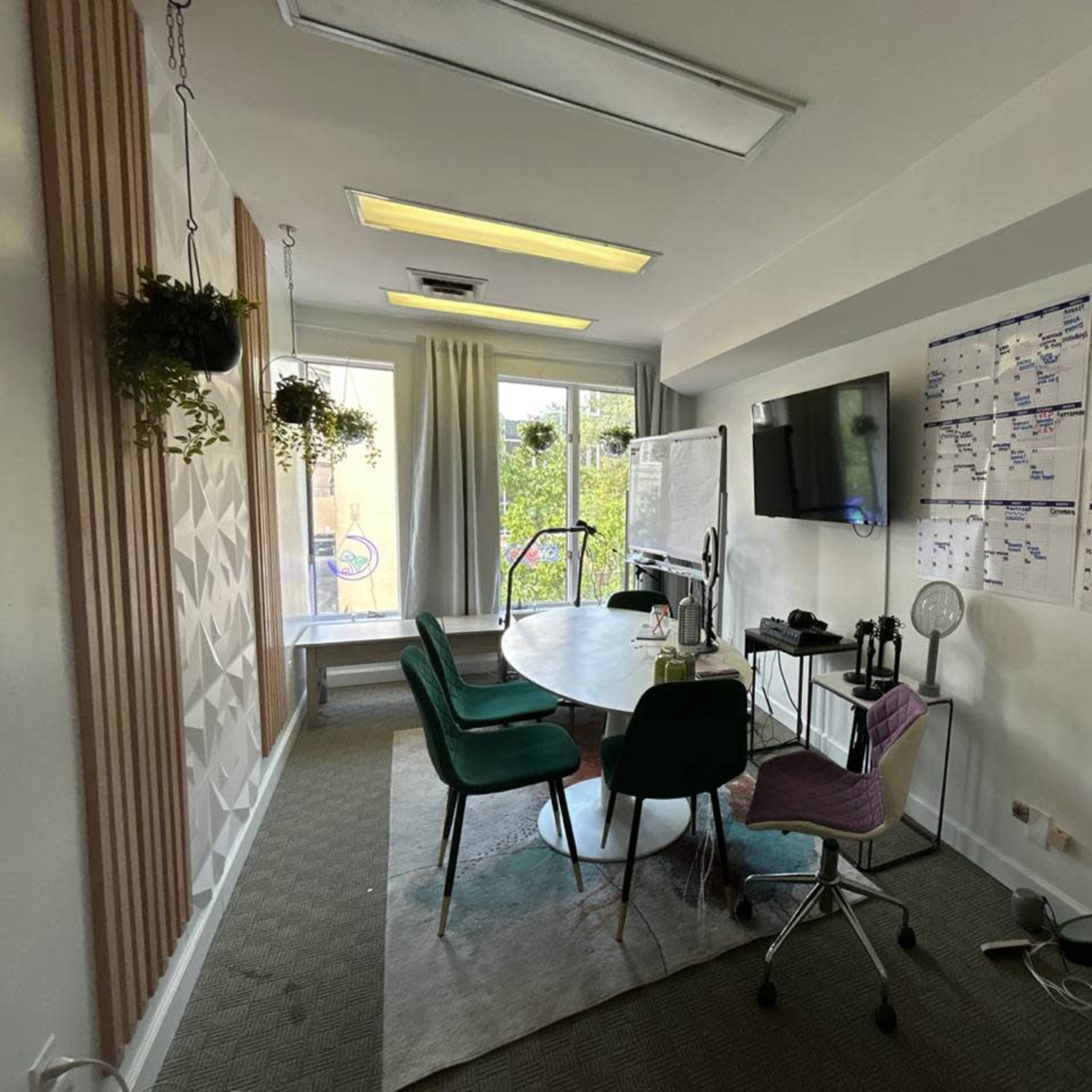 A meeting room with a round table, green chairs, a large wall-mounted screen, and a calendar on the wall, surrounded by windows and plant decor.