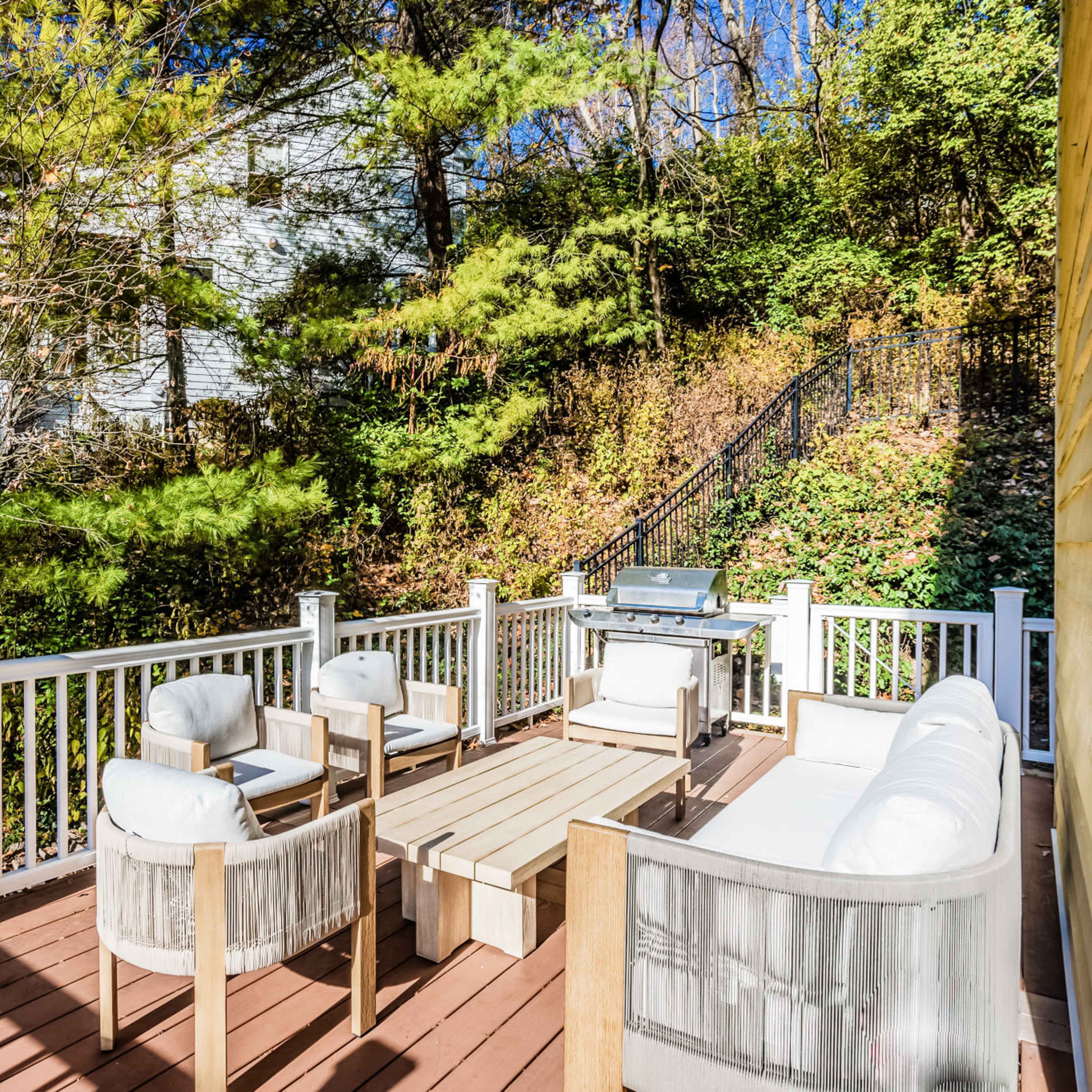A wooden deck features a seating area with two chairs, a sofa, and a low table, alongside a grill and surrounded by greenery.