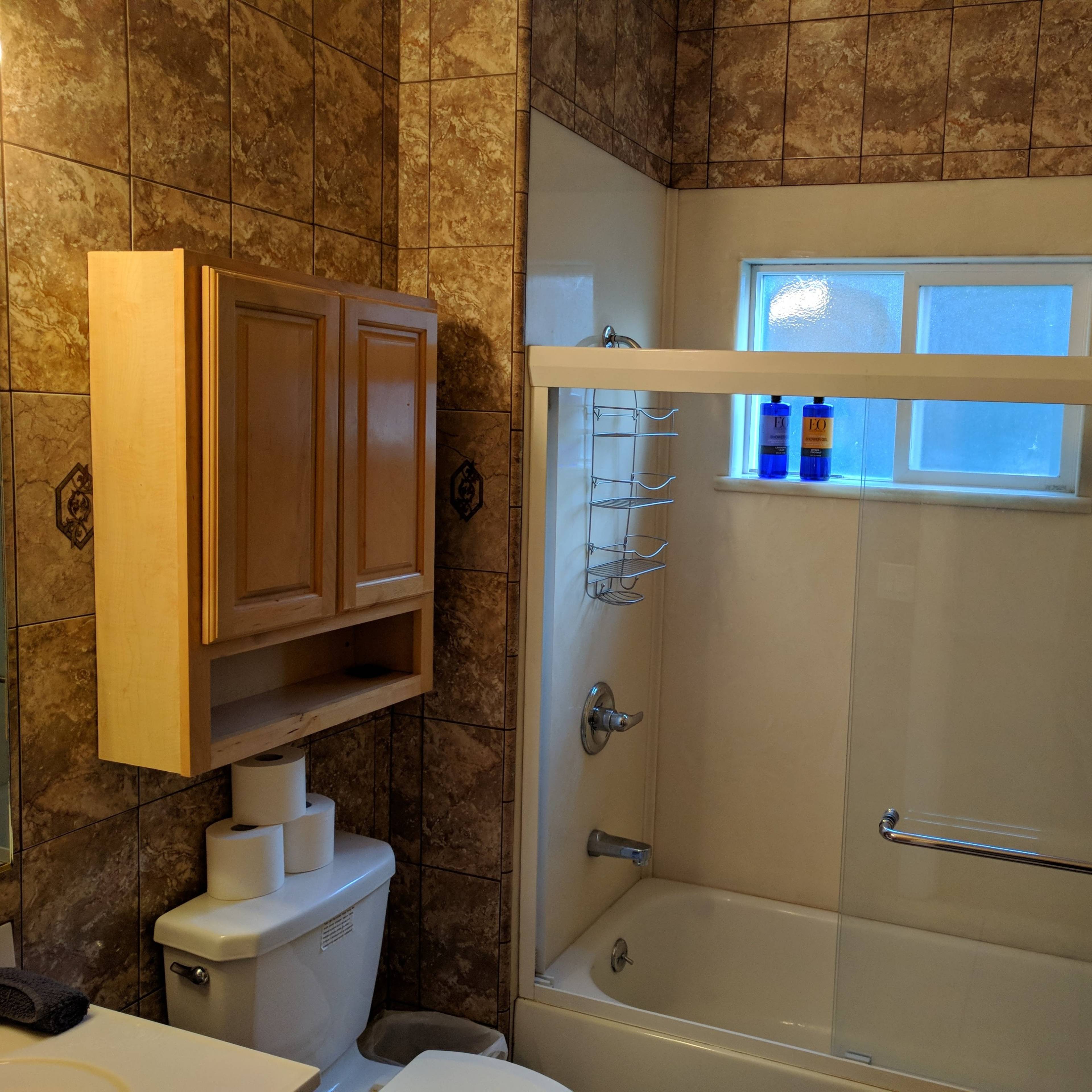 A bathroom with beige tiled walls, a white bathtub with a shower, and a wooden cabinet above a toilet.