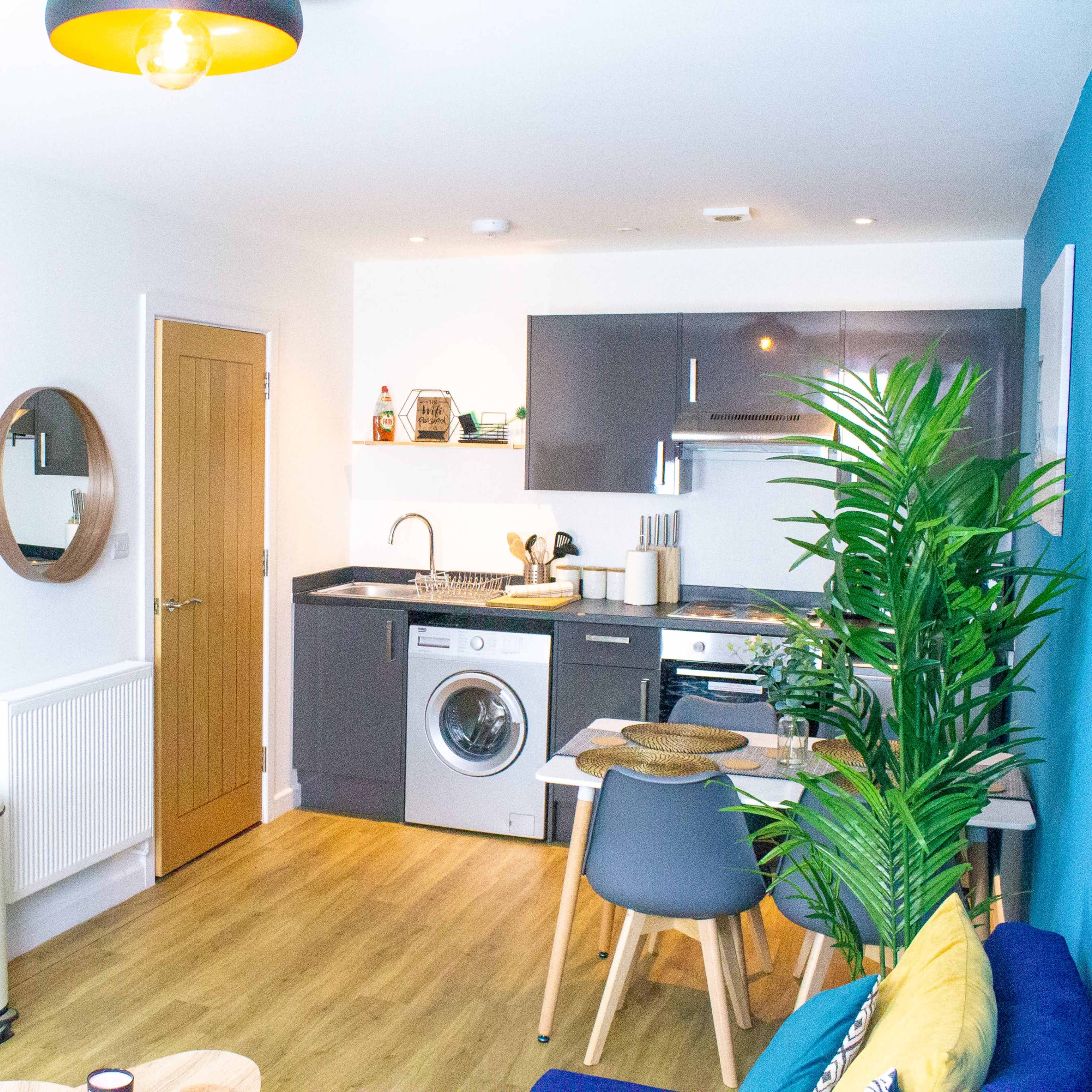 A modern, open-plan living space featuring a small kitchenette, a washing machine, and a seating area with a blue sofa and a round coffee table.