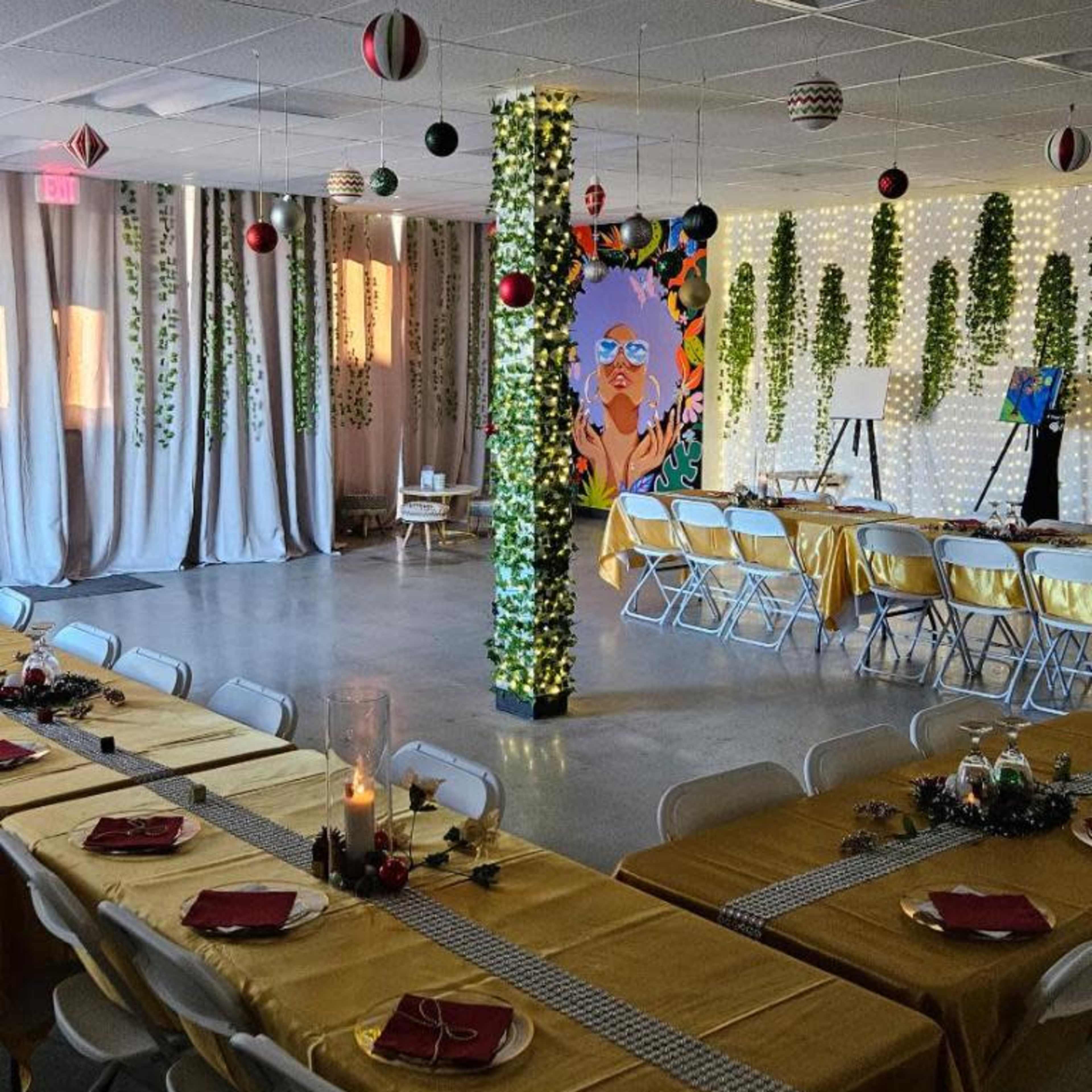 A decorated indoor event space featuring rows of tables with yellow tablecloths, hanging ornaments, and greenery on the walls and pillars.