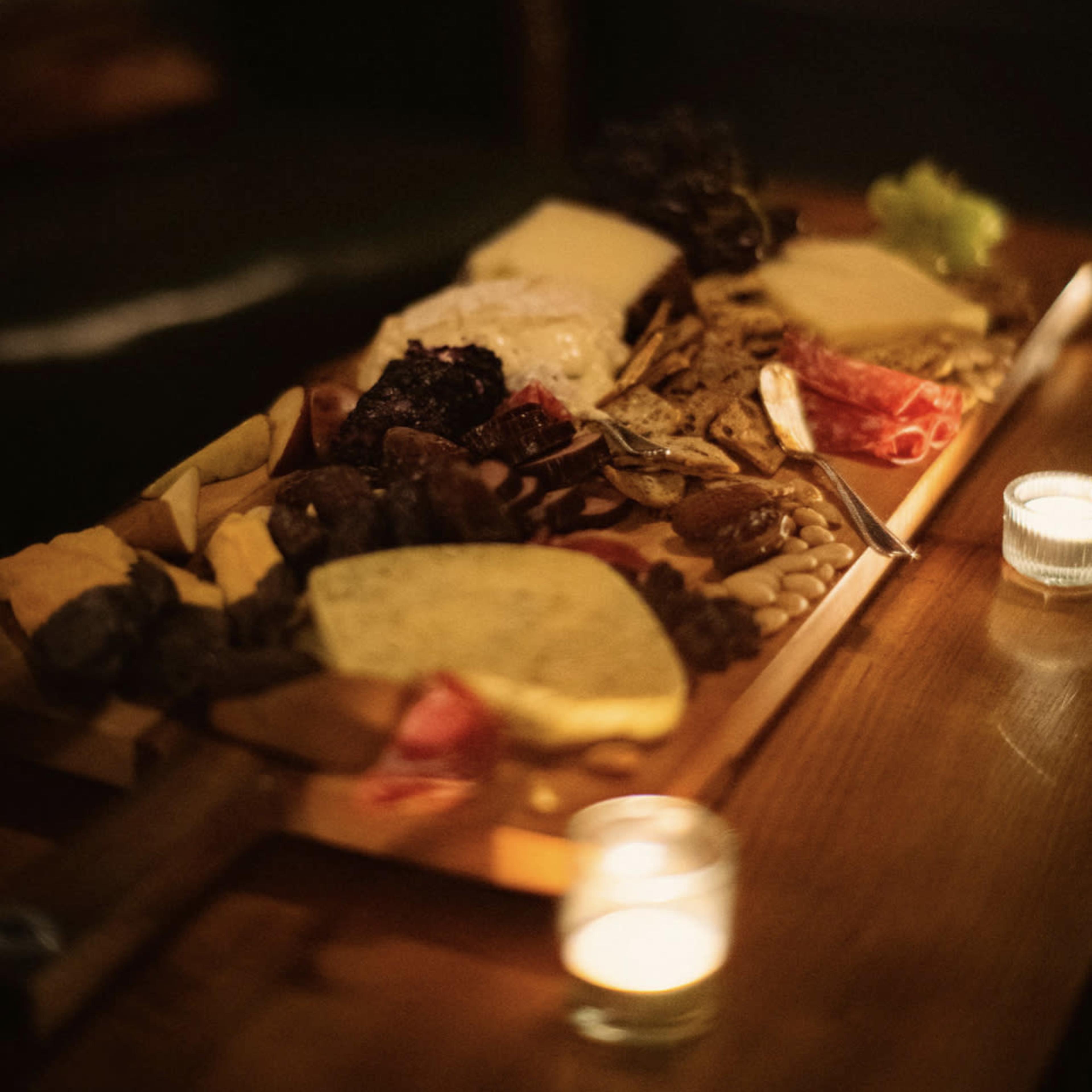 A wooden platter is filled with various cheeses, cured meats, dried fruits, and nuts, accompanied by two small candles nearby.