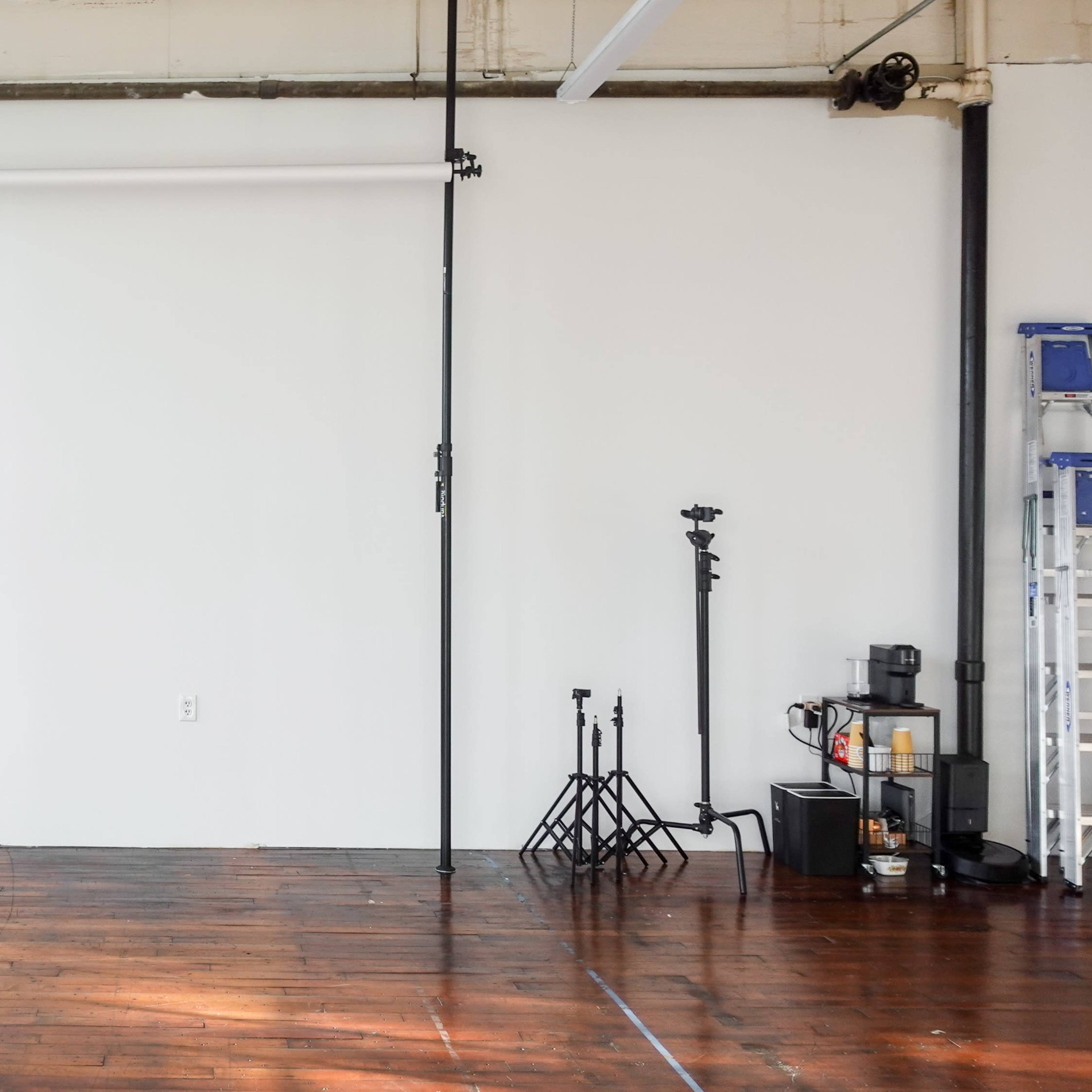 An empty studio space with wooden flooring, a white wall, and various equipment including a fan and light stands against the walls.