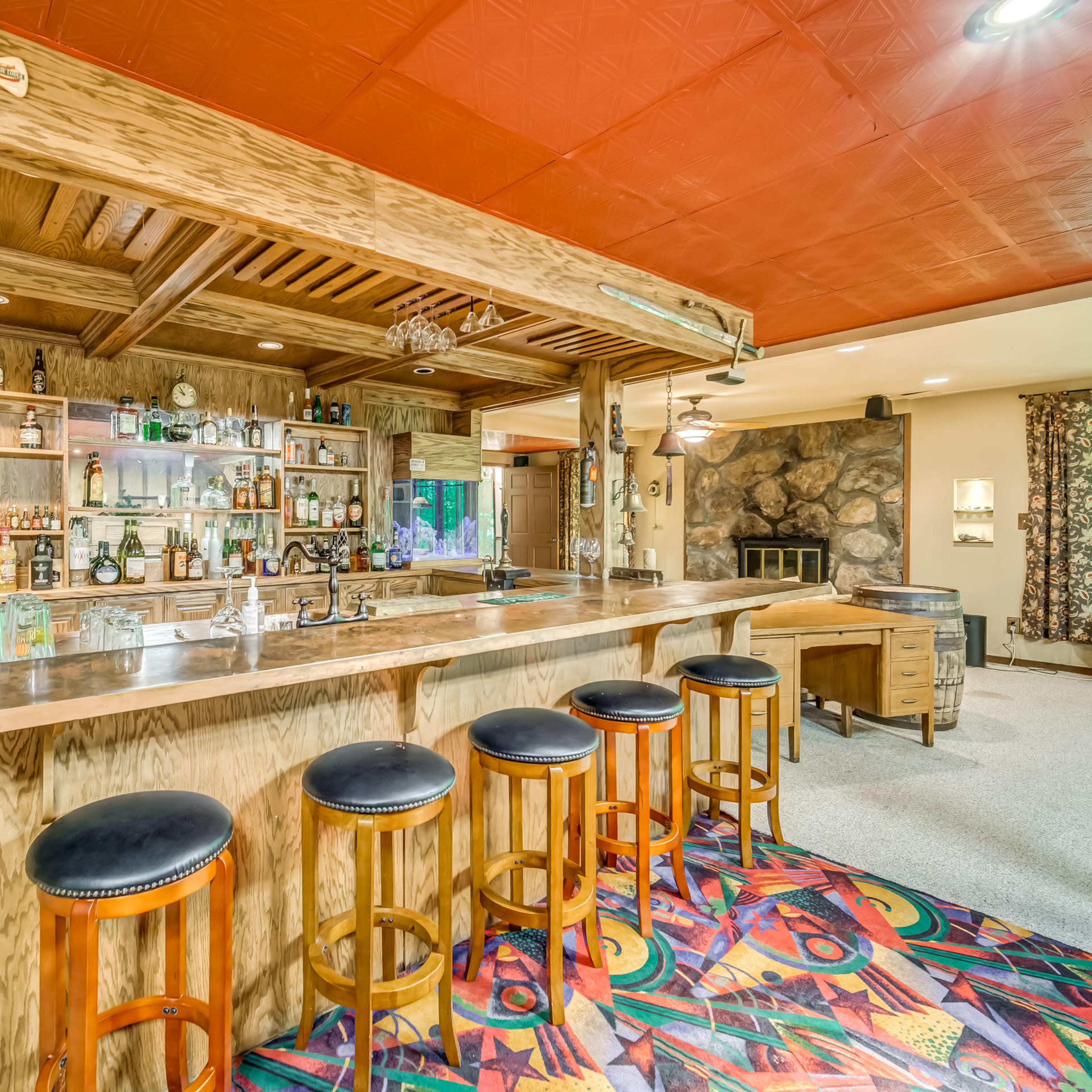 A wooden bar area with high stools, shelves of alcohol, and a stone fireplace in a lounge setting.