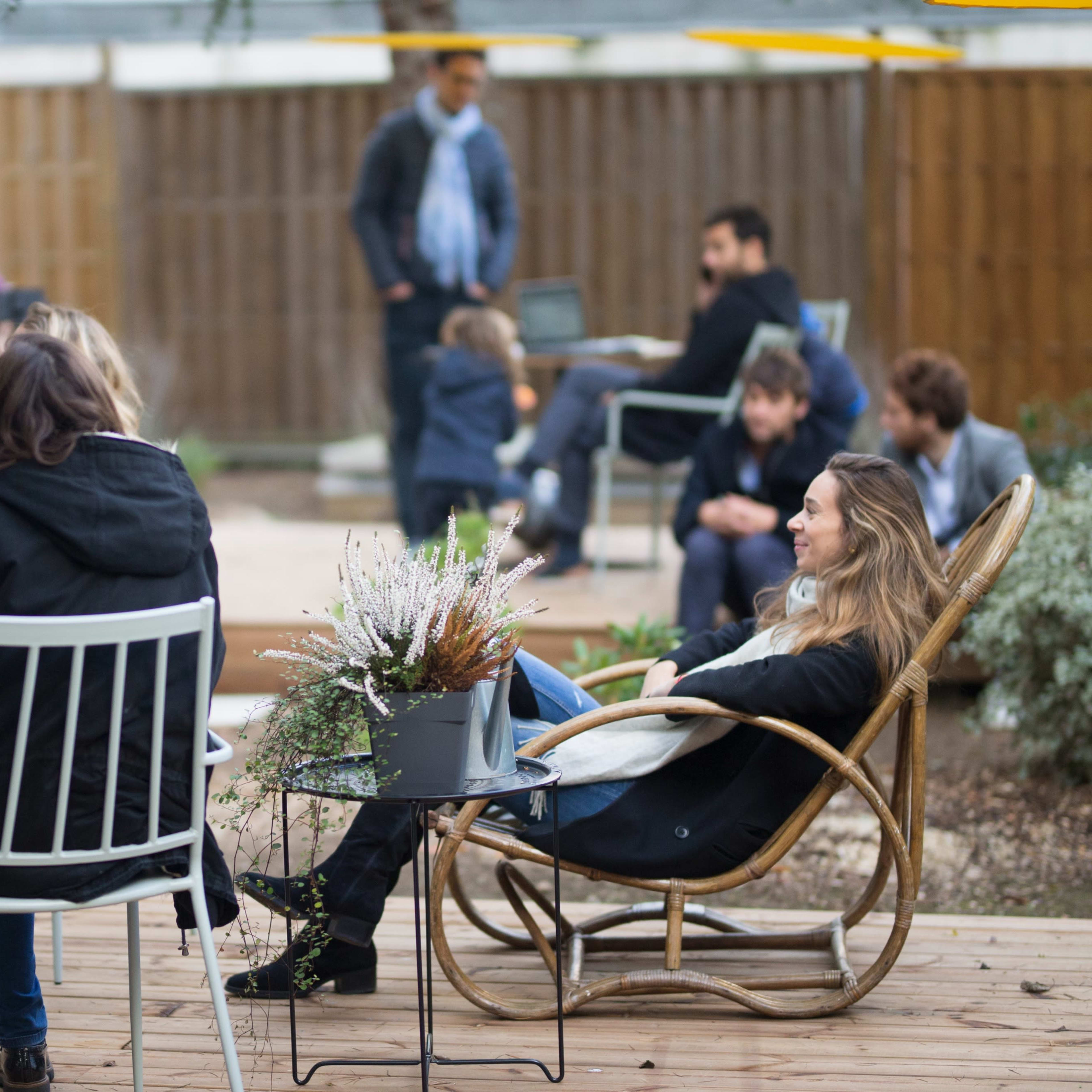 A group of people are gathered outdoors, some seated on chairs and others standing, in a casual social setting with greenery and a wooden deck.