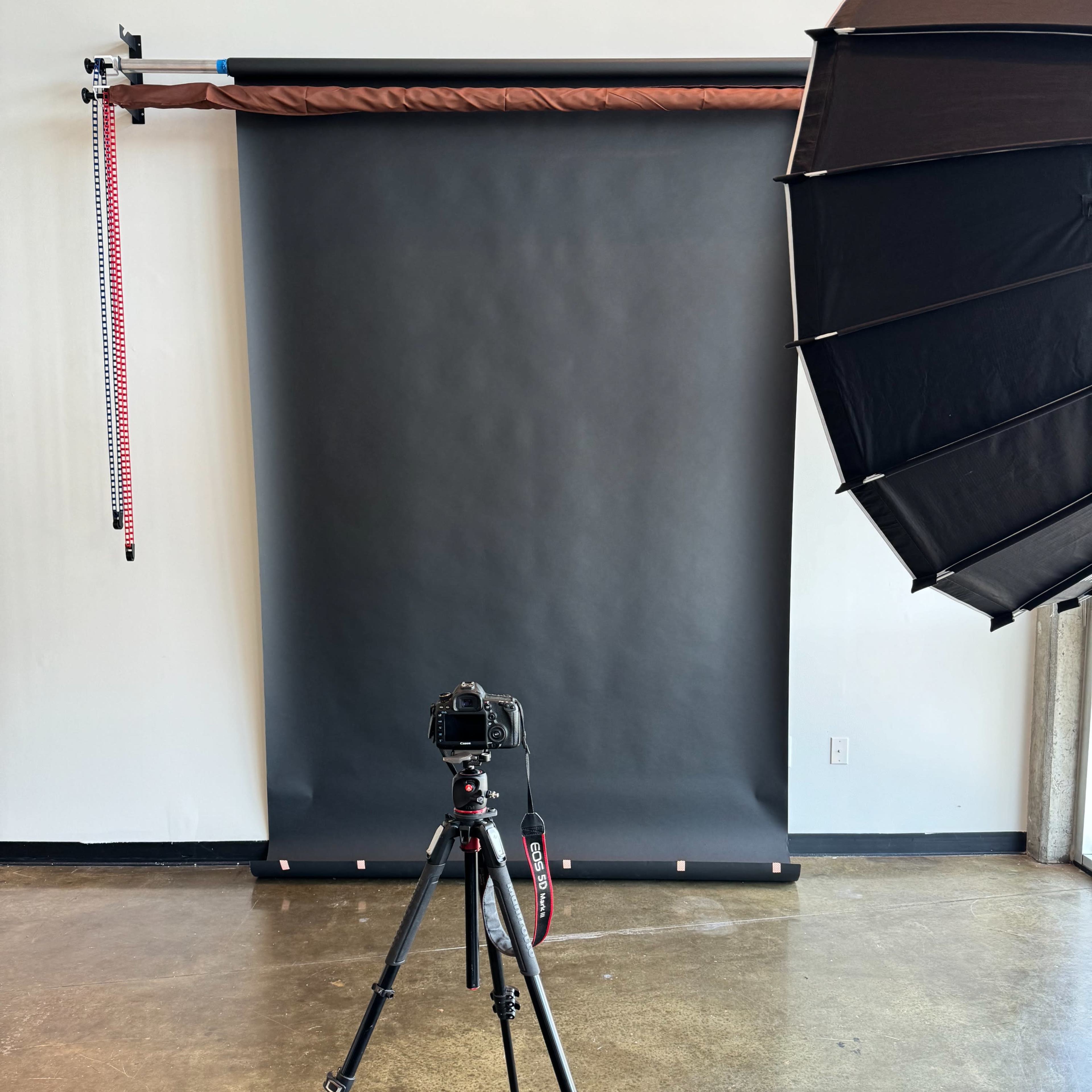 A camera mounted on a tripod is positioned in front of a dark backdrop with a softbox lighting setup to the side.
