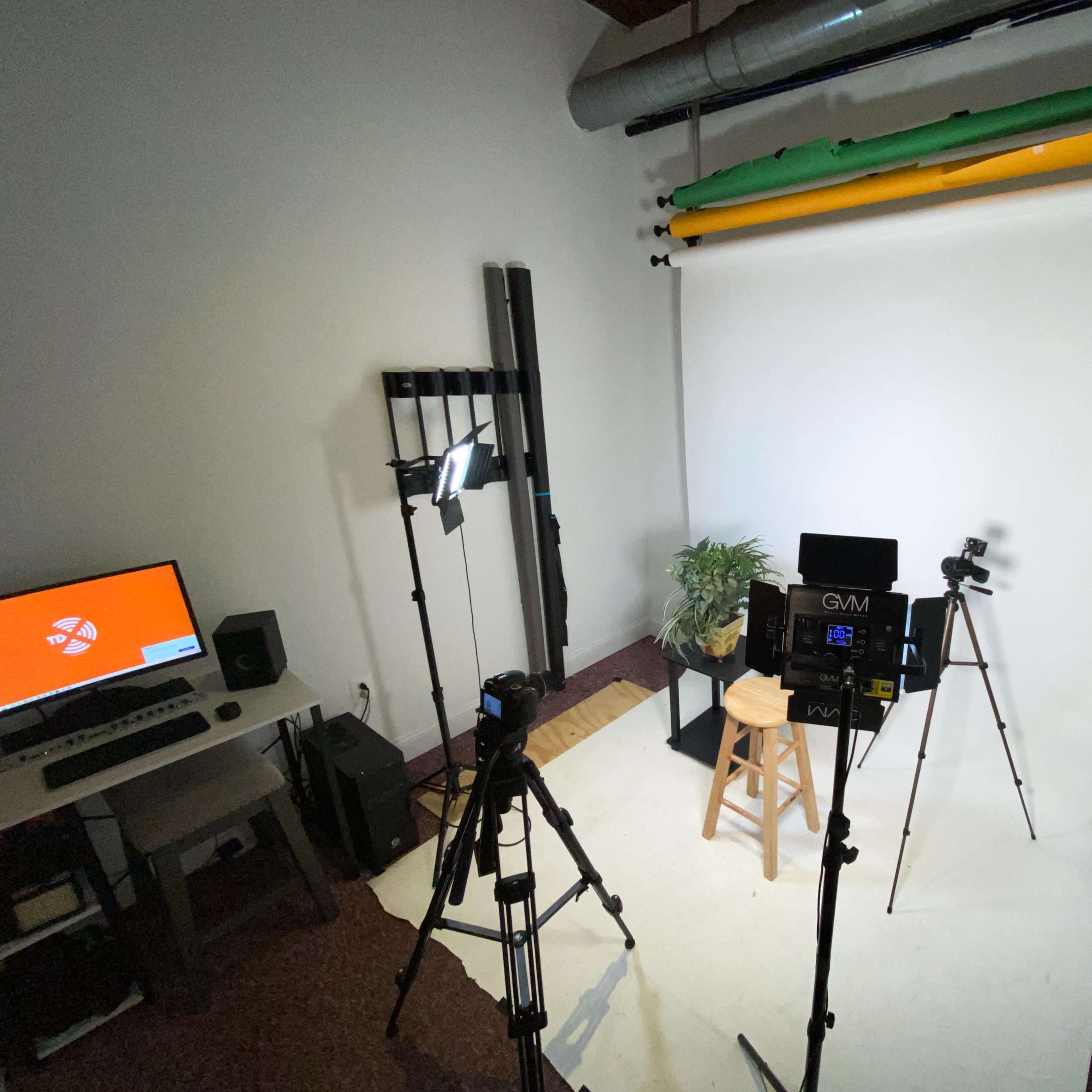 A photography studio setup with a white backdrop, studio lights, cameras on tripods, and a stool for a subject.