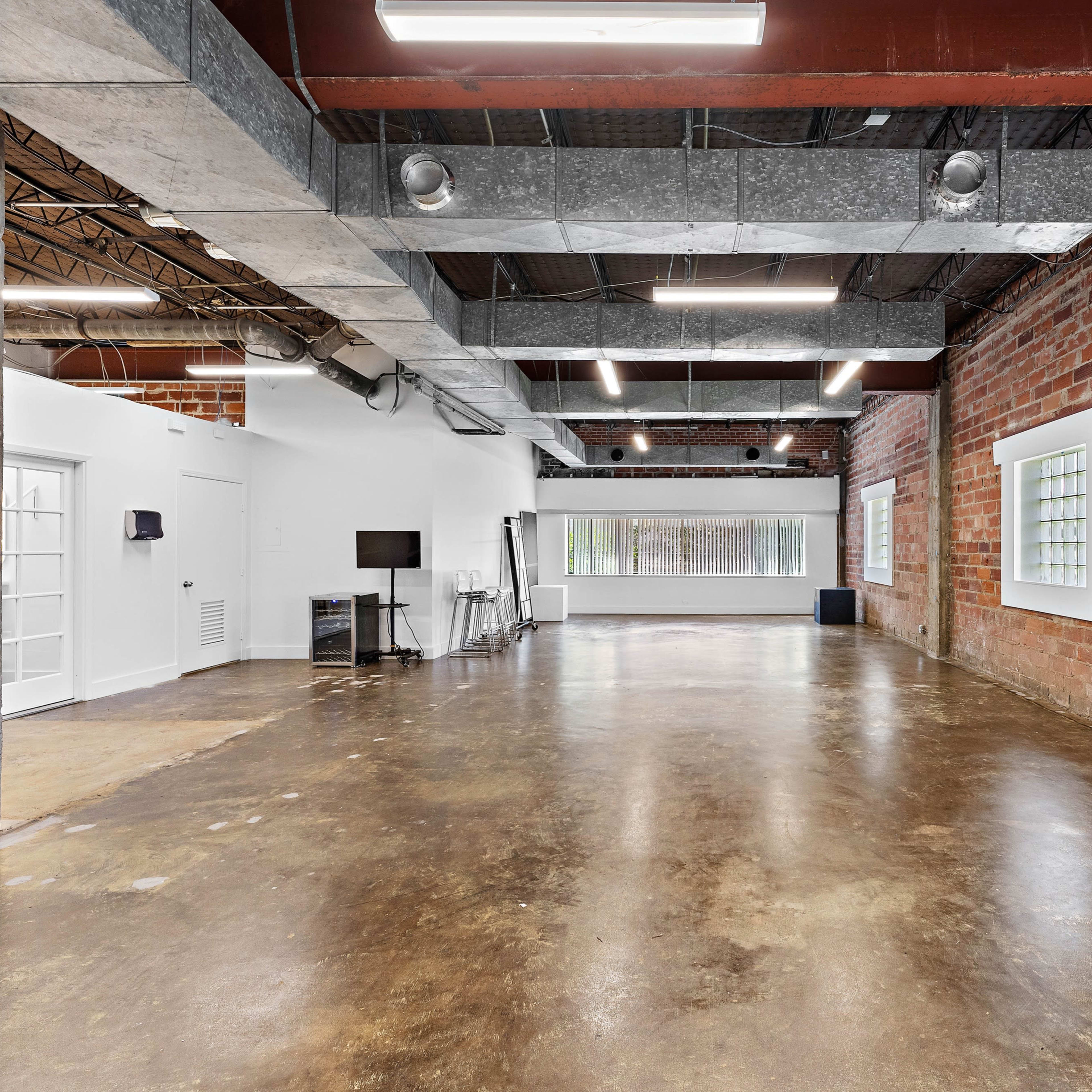 An open, industrial-style interior space with exposed brick walls, polished concrete flooring, and large windows allowing natural light.