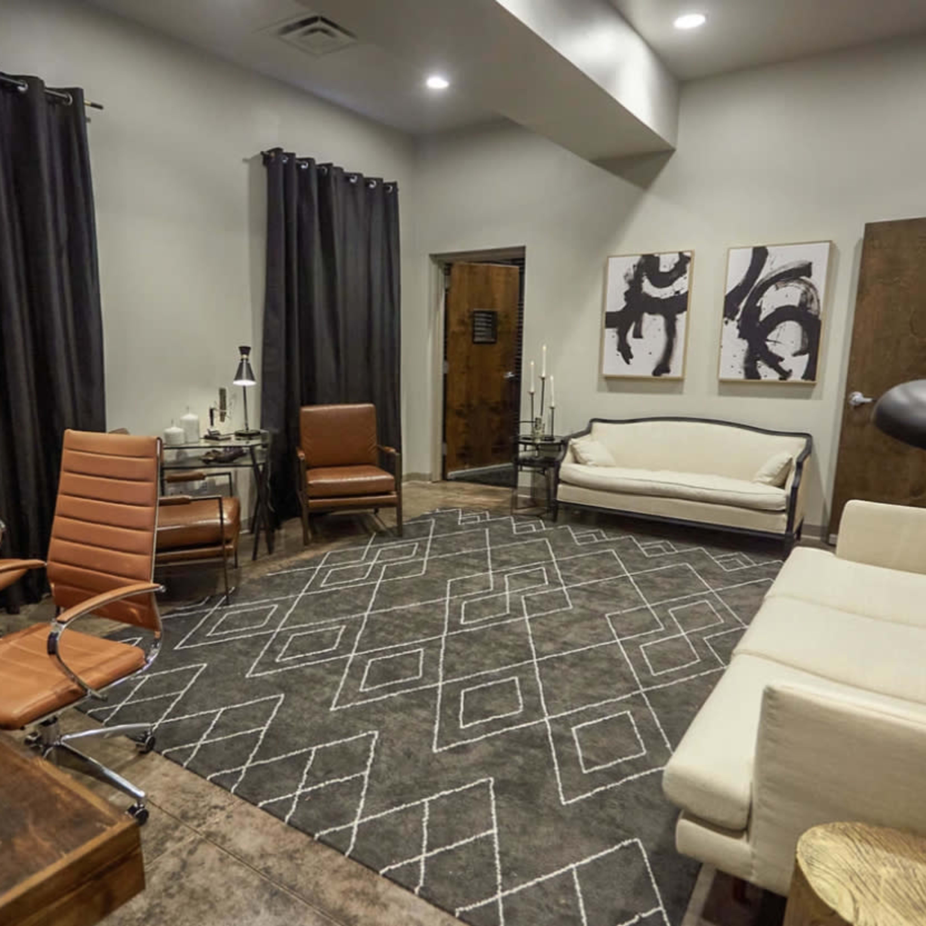 A modern office space featuring a large area rug, three chairs, a sofa, a wooden desk, and two abstract artworks on the wall.