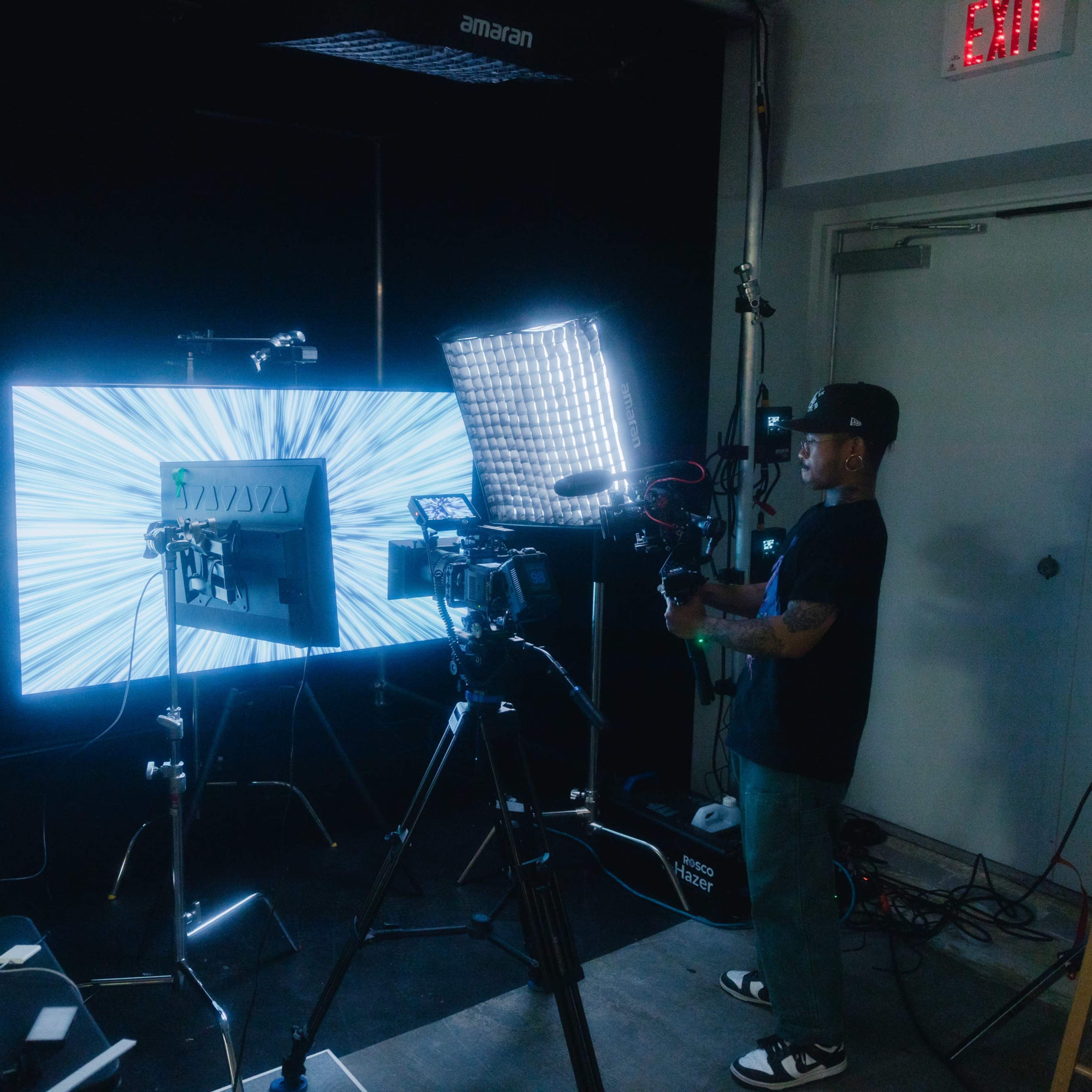 A person operates a camera setup in front of a brightly lit screen displaying a dynamic visual effect.