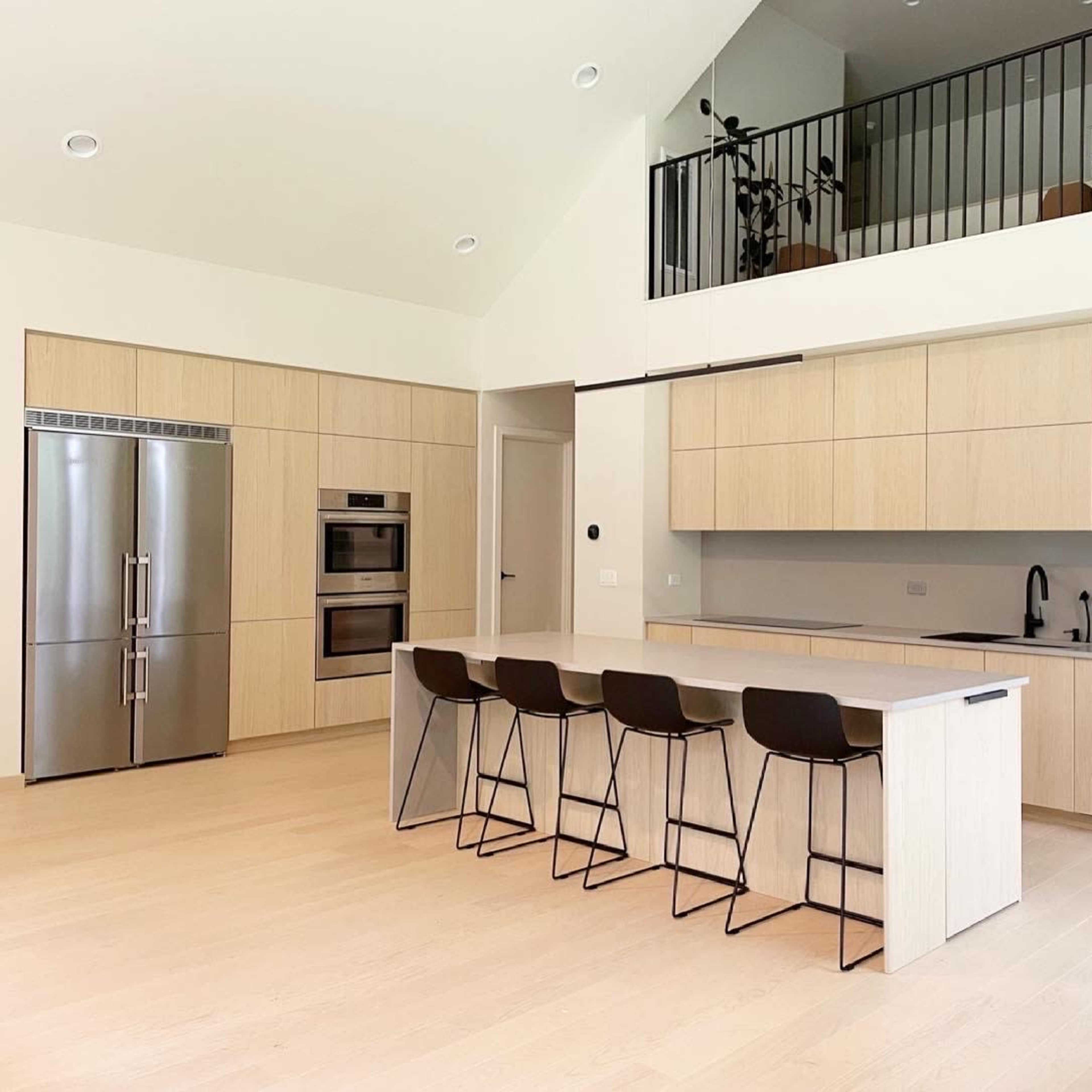 A modern kitchen with light wood cabinetry, stainless steel appliances, and a large island with four black bar stools.