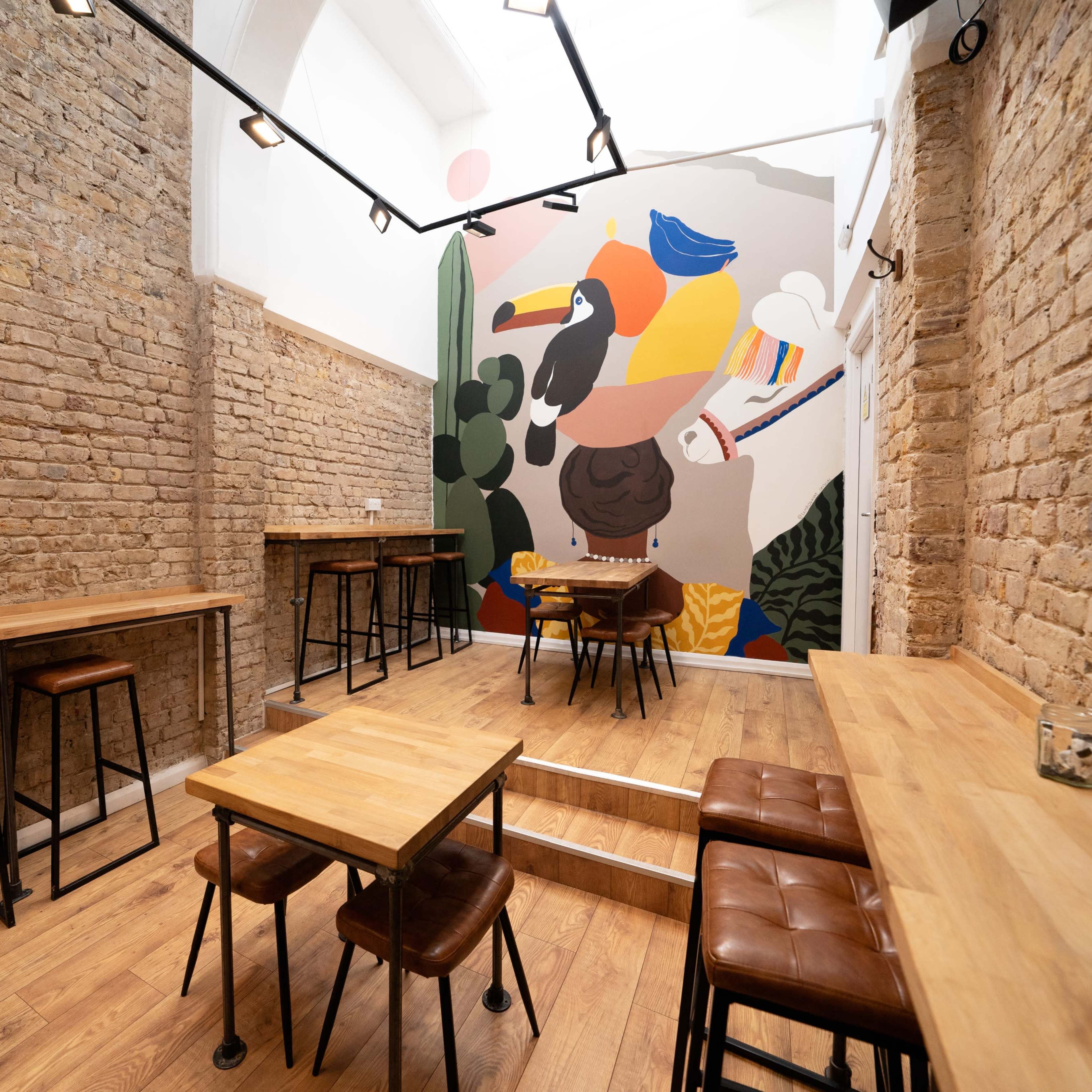 A modern cafe interior with wooden tables and stools, featuring a large colorful mural on one brick wall.