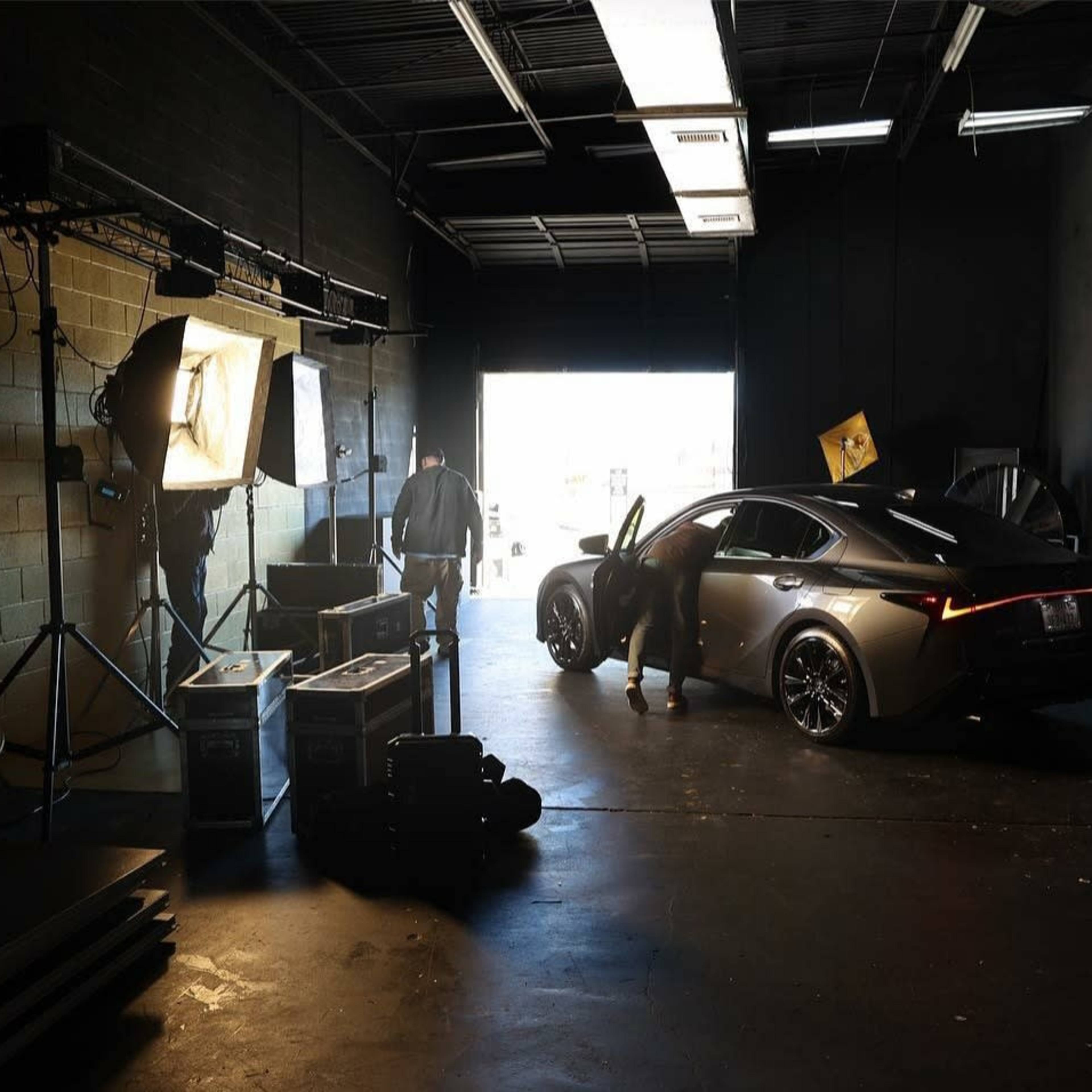 A person is unloading a car from a dimly lit garage, with studio lighting equipment and cases visible in the background.
