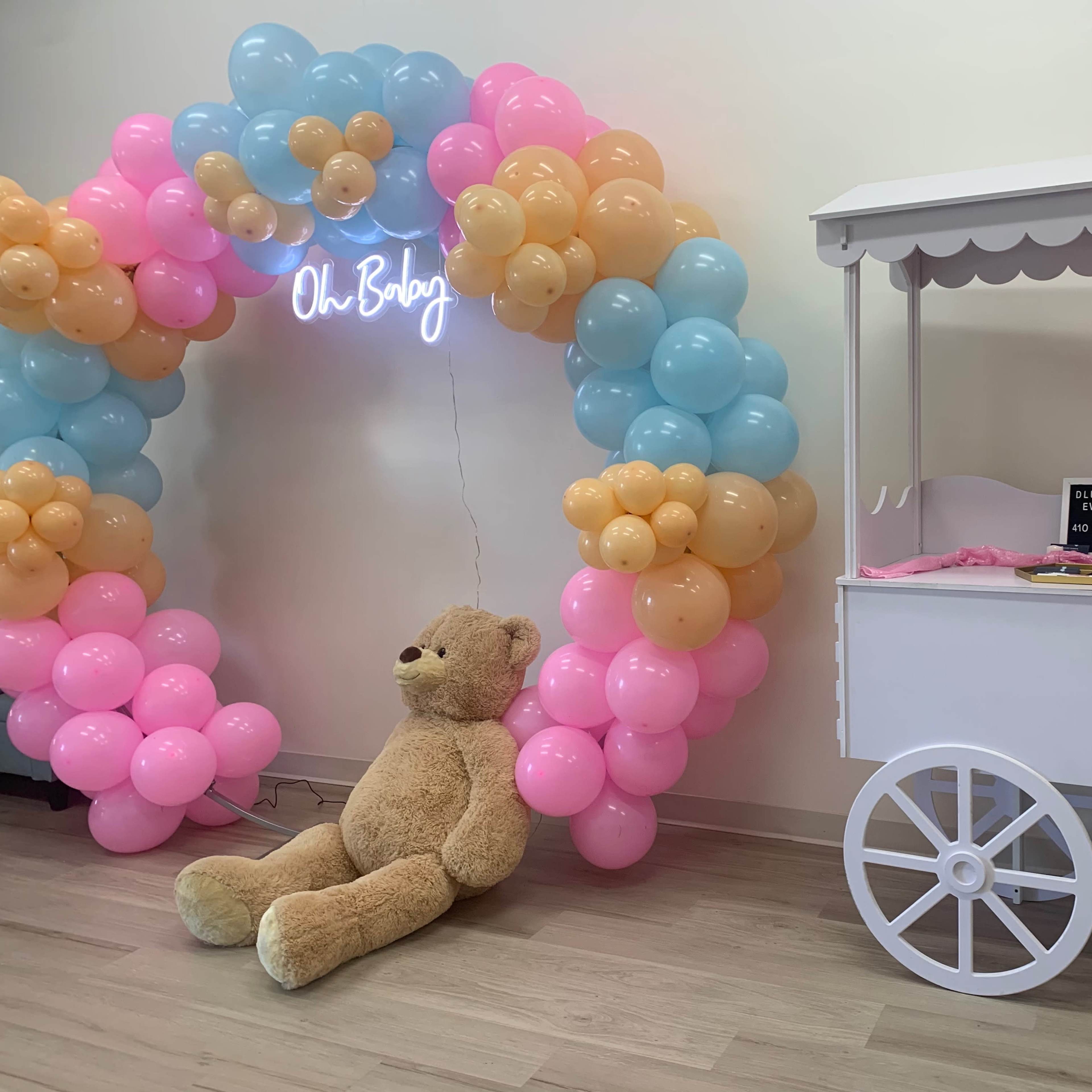 A colorful balloon arch of pink, blue, and yellow surrounds a large teddy bear sitting next to a white snack cart.