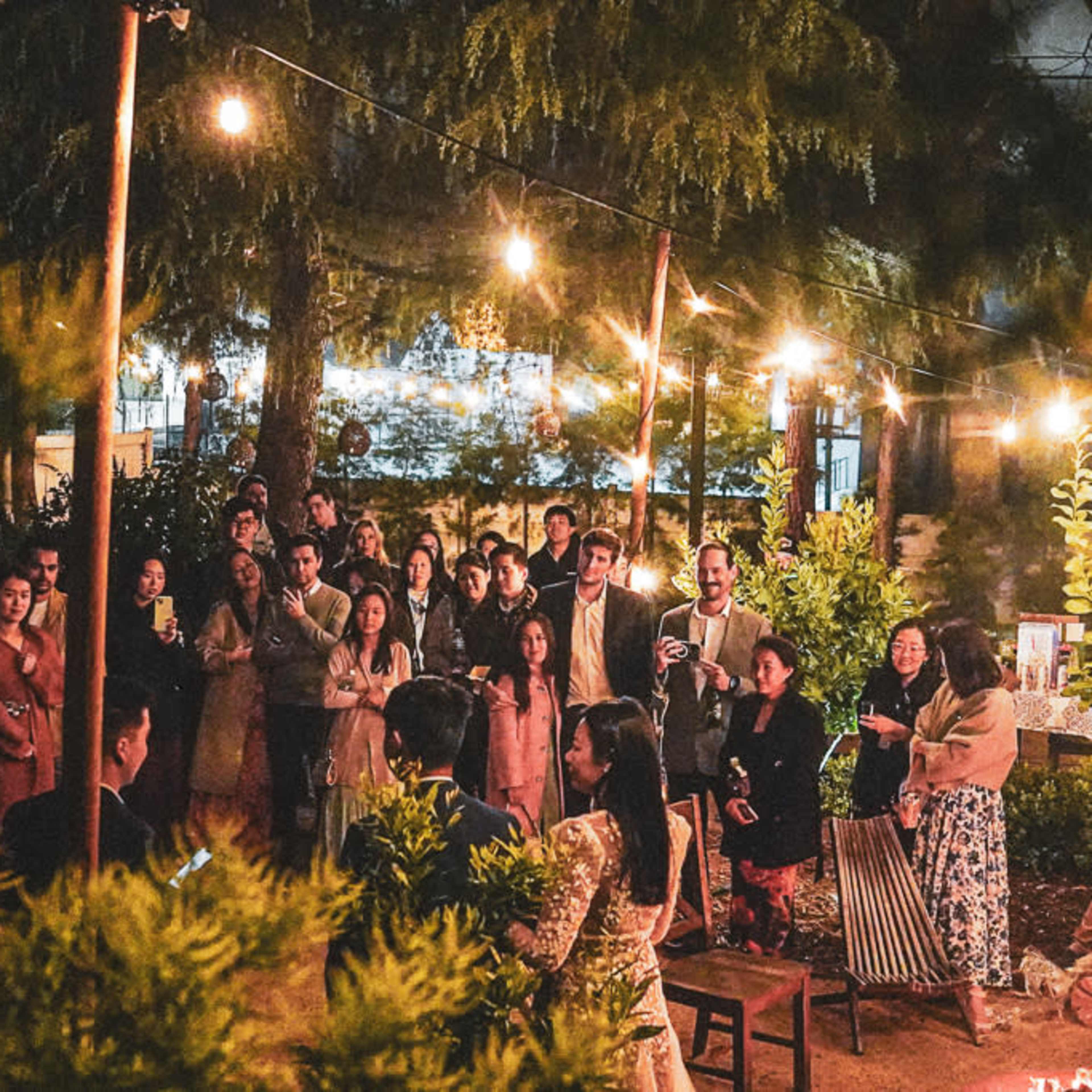 A crowd gathers around a woman speaking in a well-lit outdoor setting with string lights and trees.