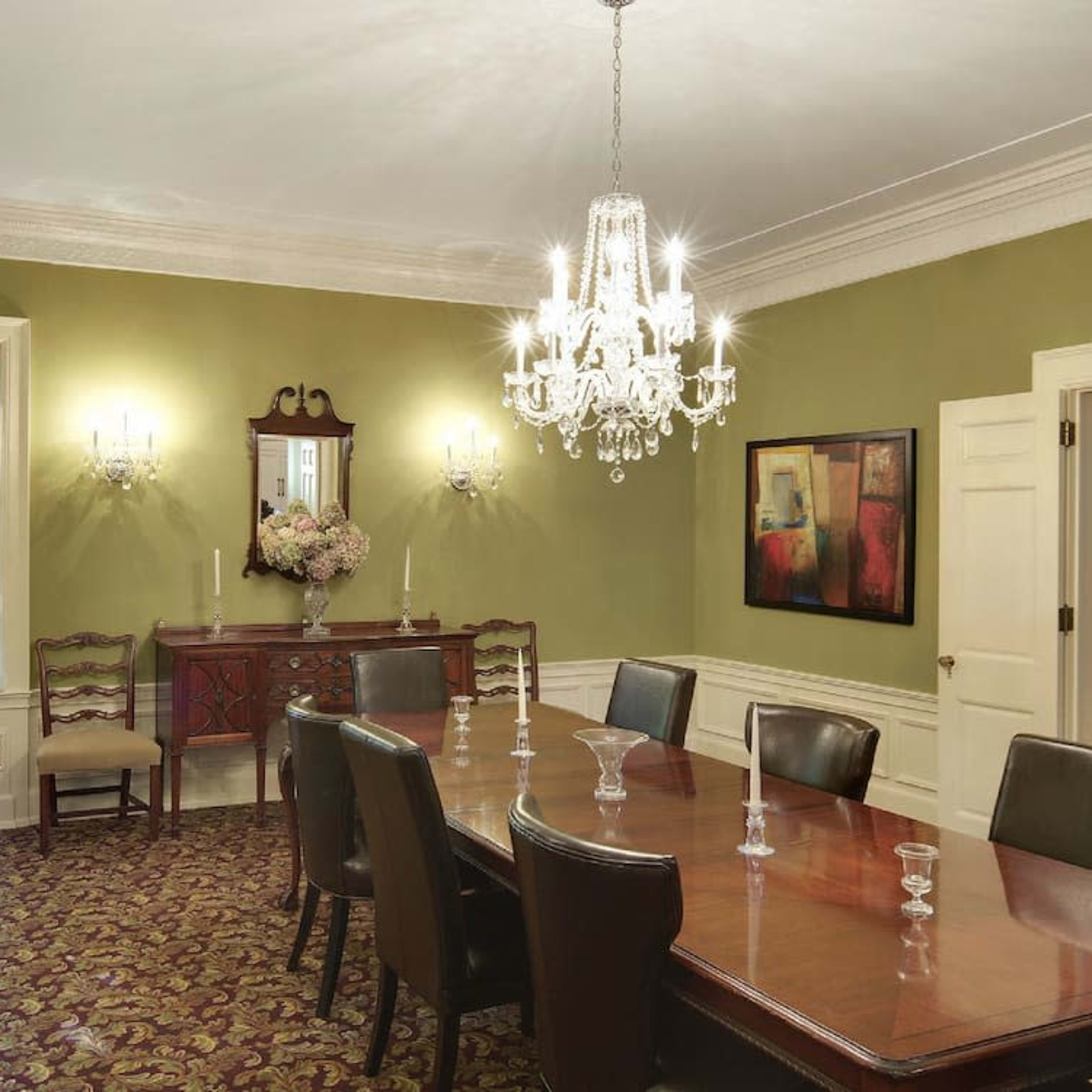 A well-furnished dining room featuring a long wooden table, chairs, a chandelier, and a sideboard against a green wall.