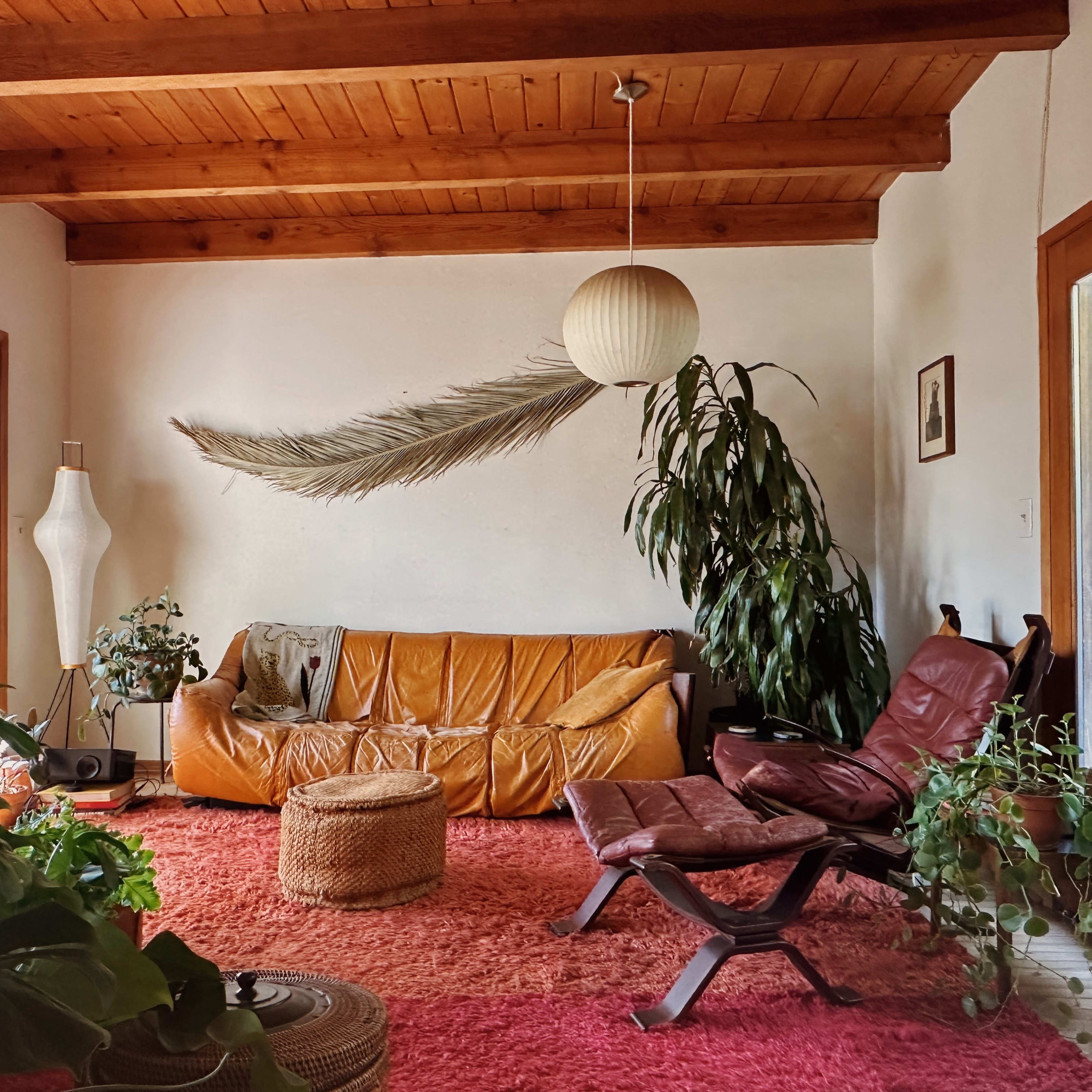 The room features a brown leather sofa, a red rug, a woven ottoman, and large windows surrounded by greenery.