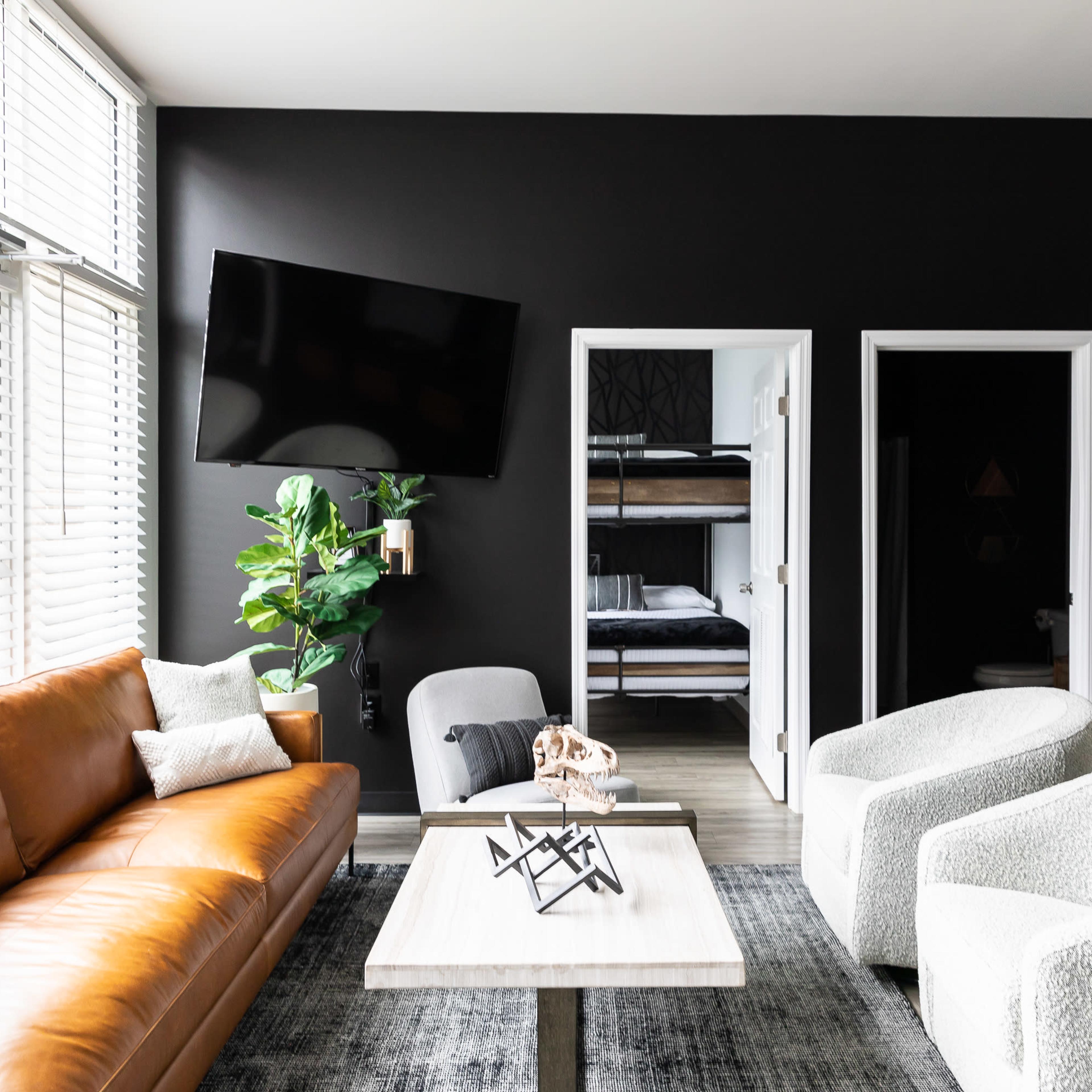 A modern living room features a brown leather sofa, two white armchairs, a wooden coffee table, and a large television mounted on a dark wall, with a potted plant beside a doorway leading to a bunk bedroom.